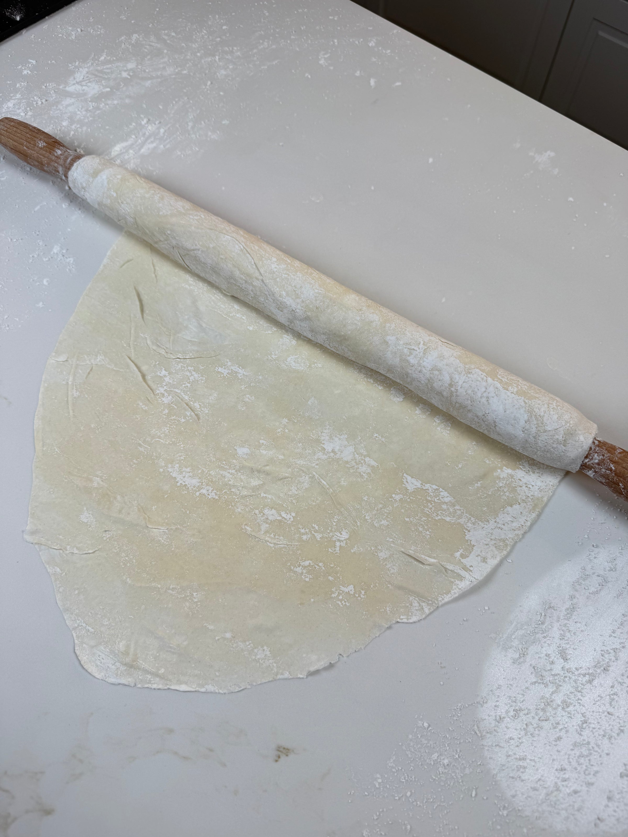 A wooden rolling pin is rolling out thin, unevenly shaped dough on a white, floured surface, with flour scattered around.