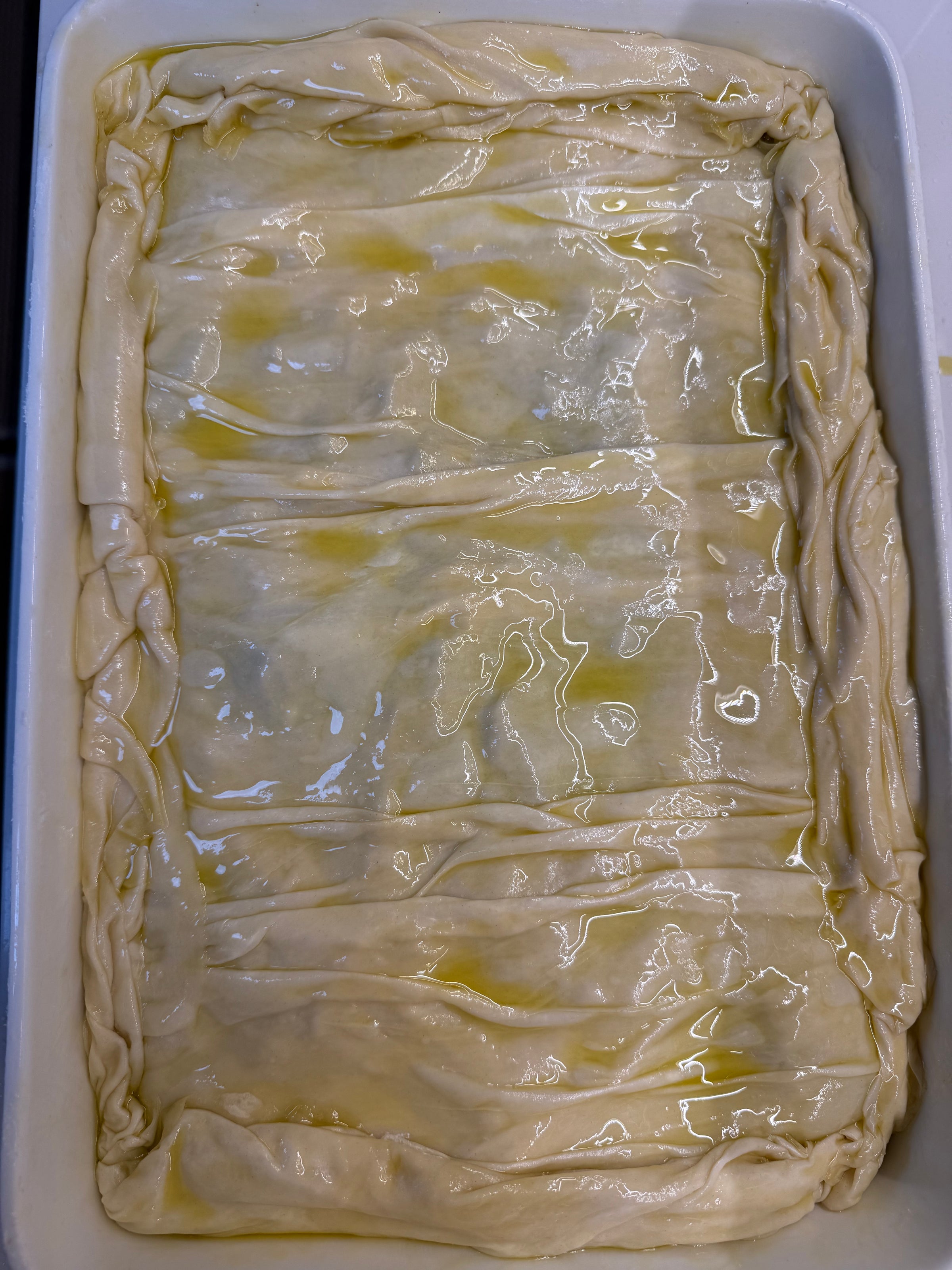 A rectangular baking dish filled with unbaked, layered phyllo dough that is brushed with oil or butter, with the edges slightly twisted and crimped around the border.