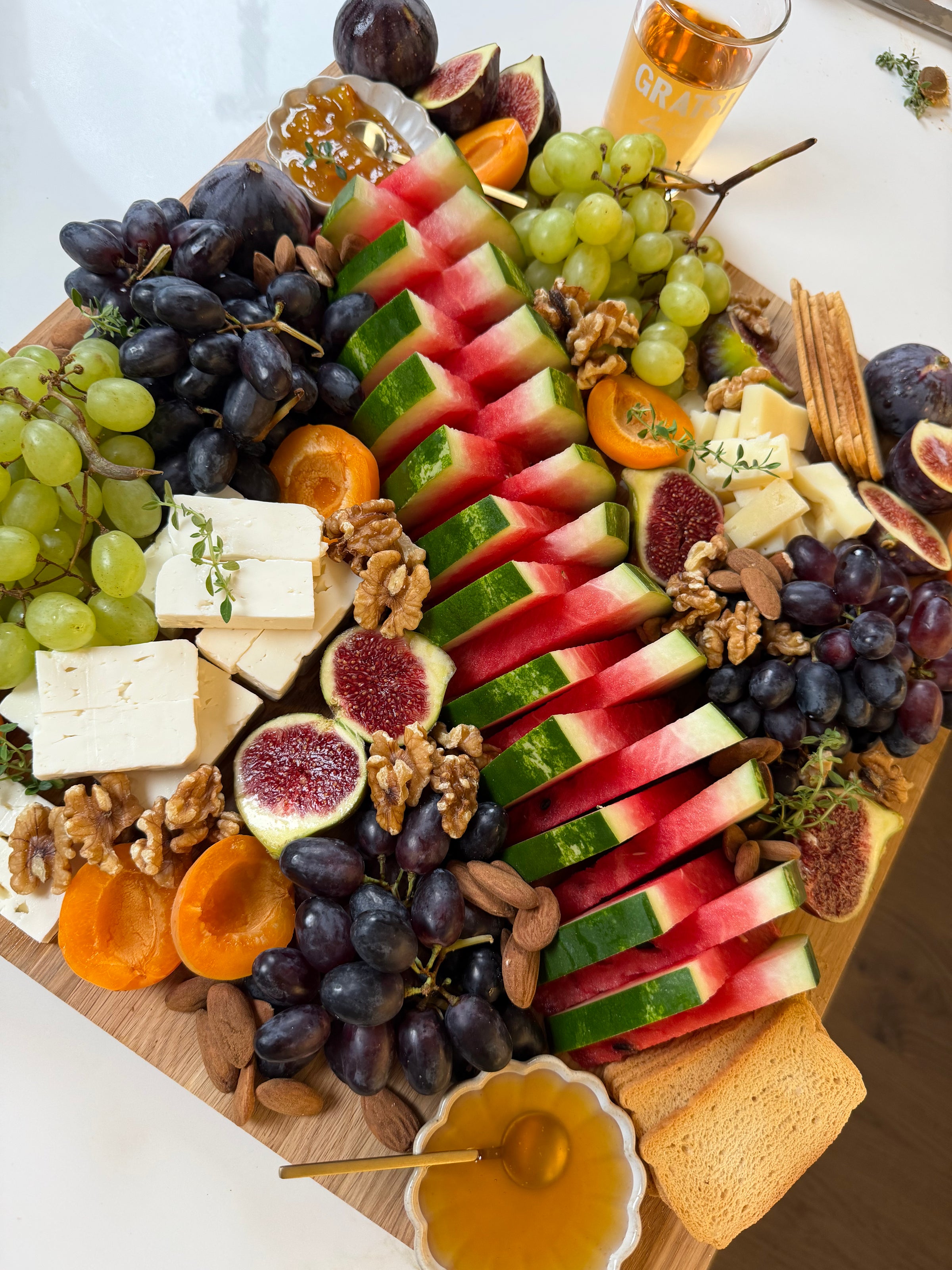 Charcuterie board with sliced watermelon, grapes, figs, apricots, nuts, feta cheese, honey, crackers, bread, and a glass of drink. The arrangement is colorful and fresh.