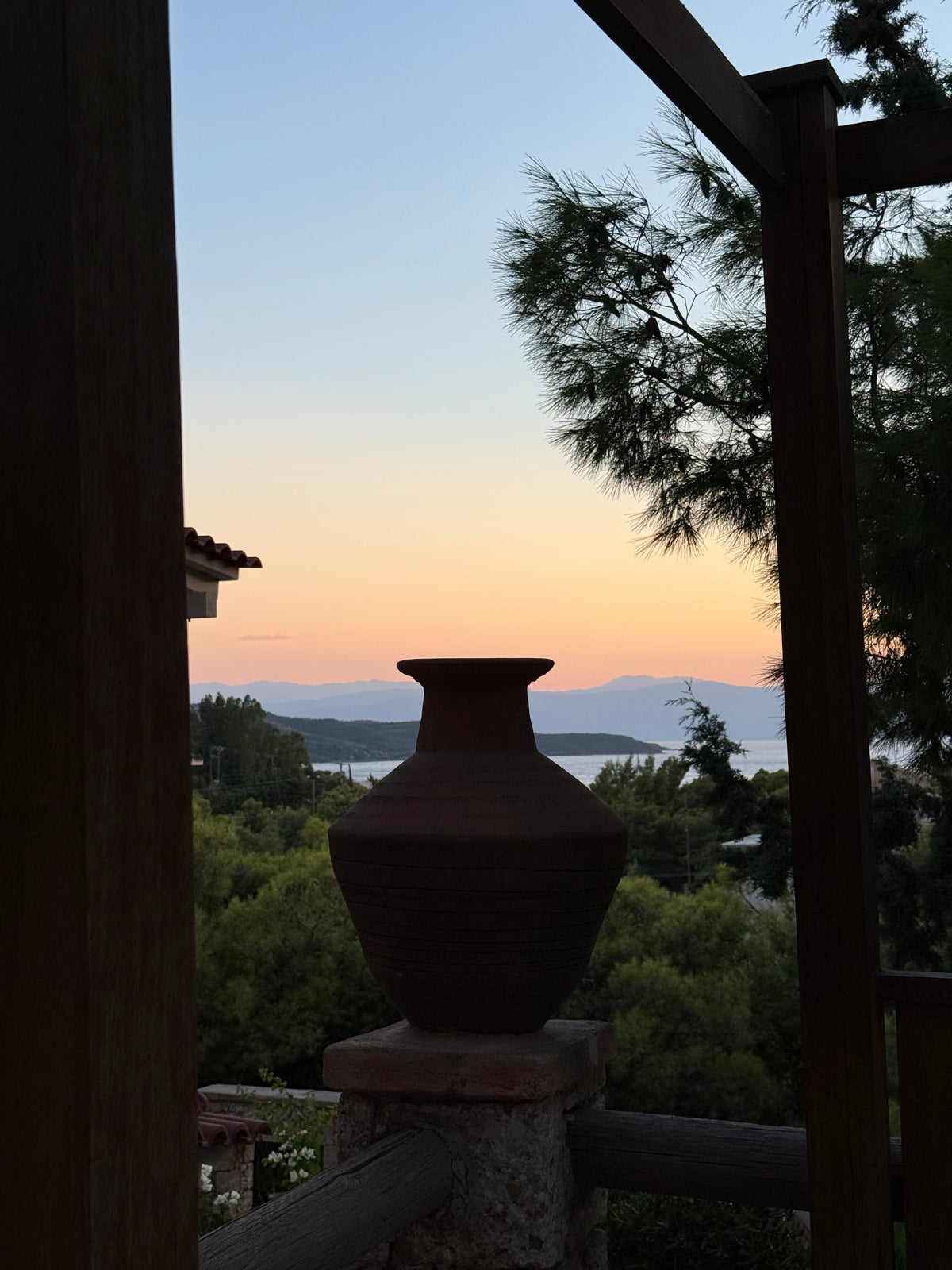 A large clay vase is silhouetted in the foreground against a scenic sunset, with trees, distant mountains, and calm water visible in the background. Wooden beams and railings frame the image.