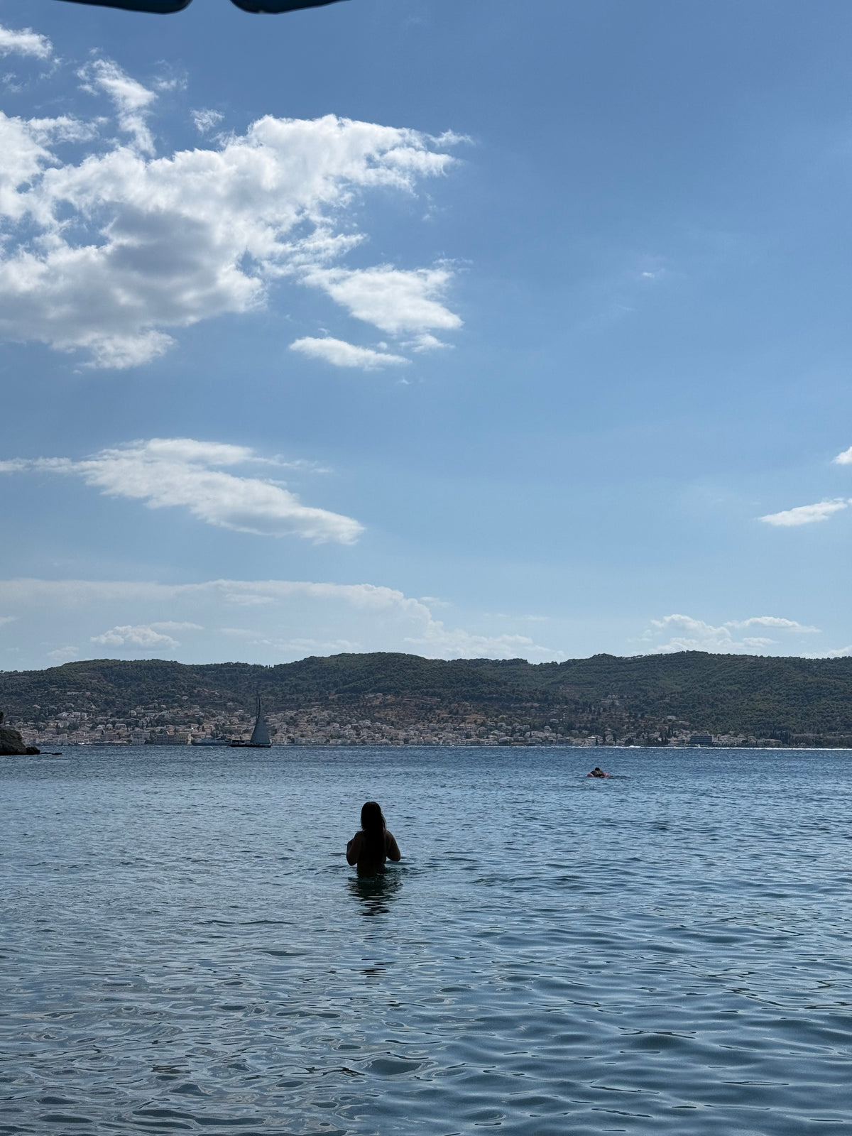 A person stands in calm water facing distant hills under a blue sky with scattered clouds. A sailboat and another person in the water are visible in the background.
