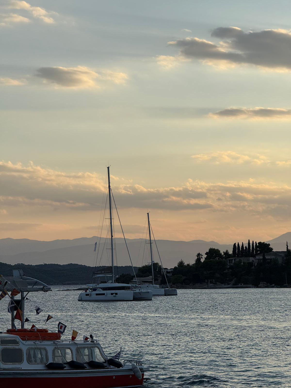Two sailboats float on calm water at sunset, with a red boat in the foreground. Trees and houses line the distant shore, and mountains appear under a sky with scattered clouds.
