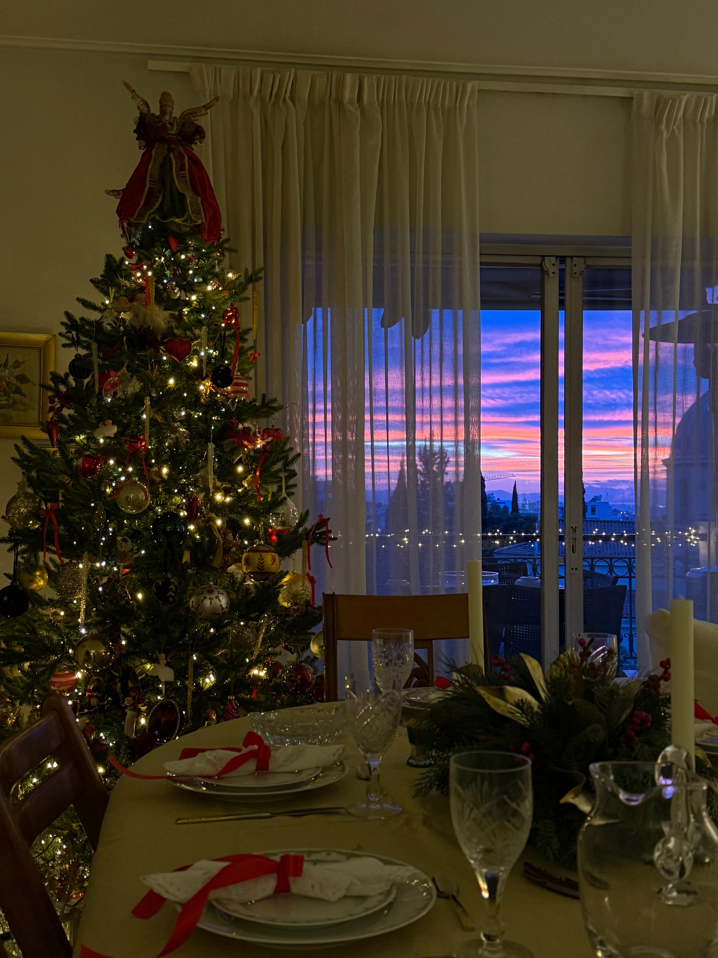 A festive dining table set for Christmas near a decorated tree, with a glowing sunset visible through glass doors in the background.