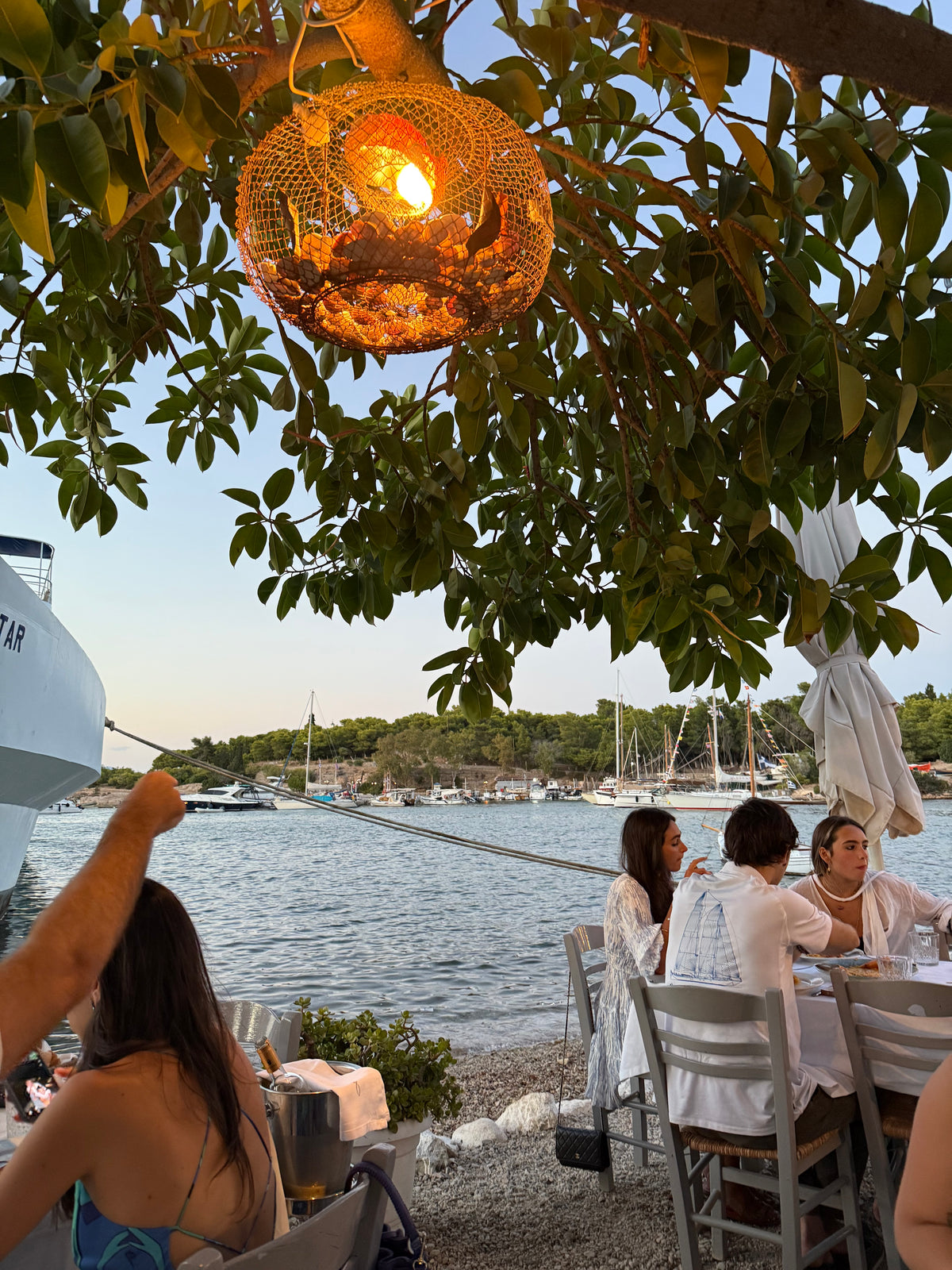 People dining at an outdoor seaside restaurant under a warm hanging light, with boats and sailboats on the calm water and trees in the background during sunset.