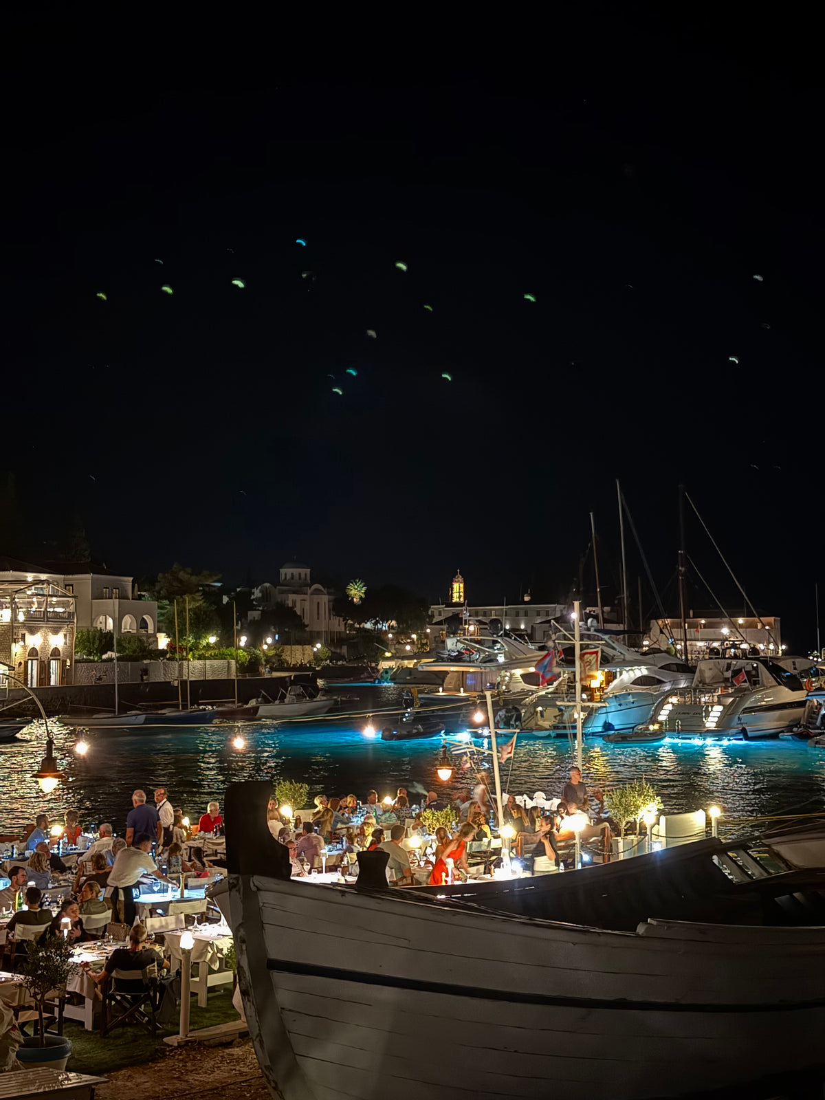 A lively outdoor waterfront restaurant at night, with people dining, boats docked nearby, and festive lights reflecting on the water.
