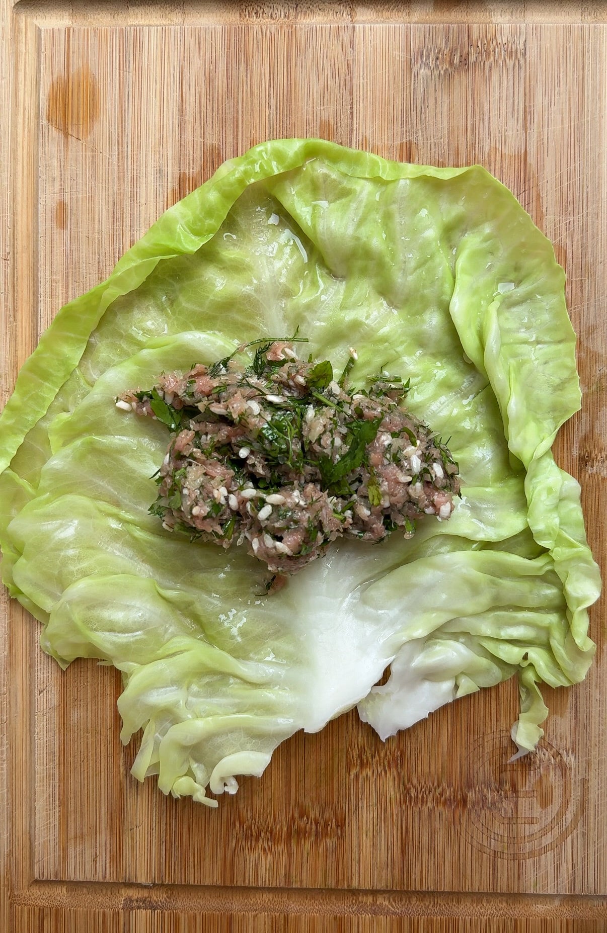 A large cabbage leaf rests on a wooden board, topped with a portion of uncooked minced meat mixed with herbs and grains, ready for wrapping.