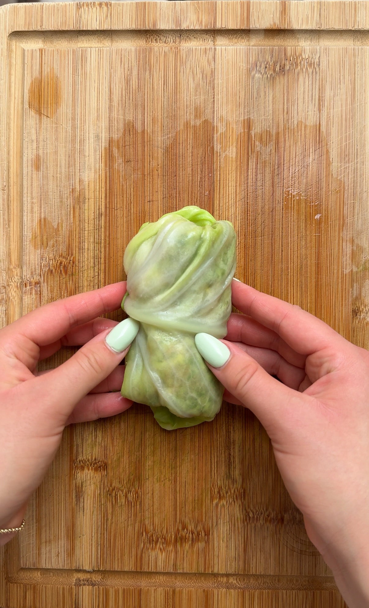Two hands with pale green nails are folding a cabbage leaf wrap on a wooden cutting board.
