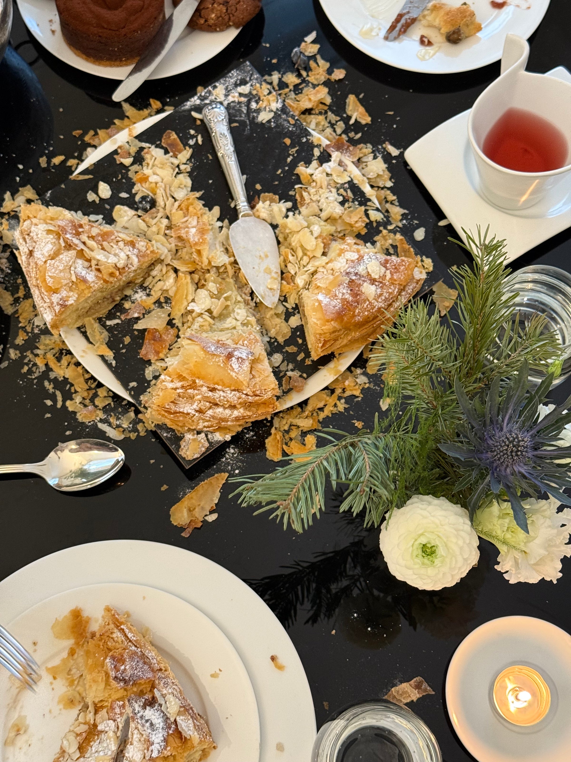 A black table with a partially eaten almond cake, crumbs scattered, a cup of tea, white flowers, a candle, and plates with cake slices. Utensils and a serving knife are also visible.