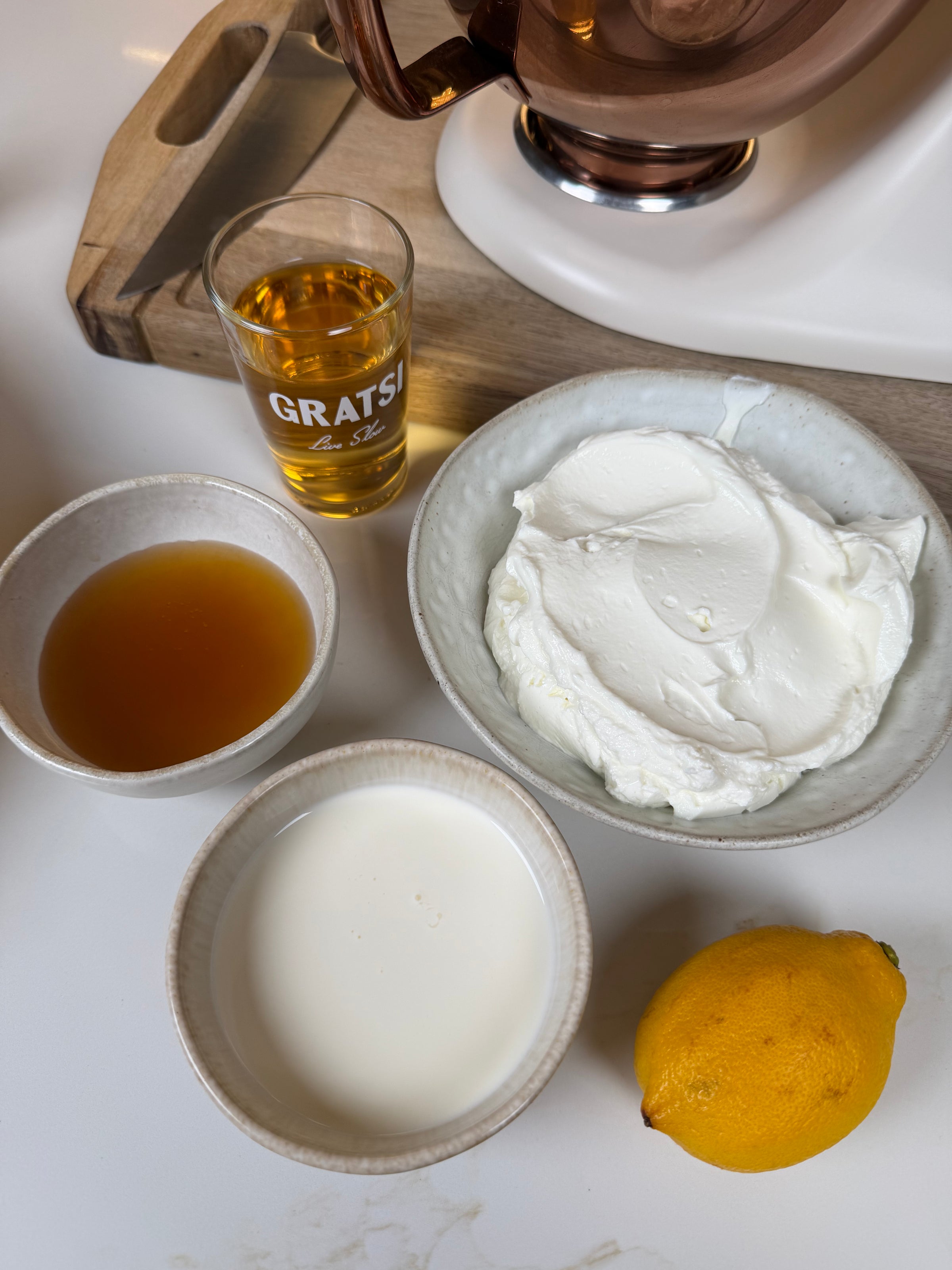 Four bowls and a glass on a counter hold honey, heavy cream, mascarpone, lemon, and a golden liquid, likely liqueur, with a wooden cutting board and a mixer in the background.