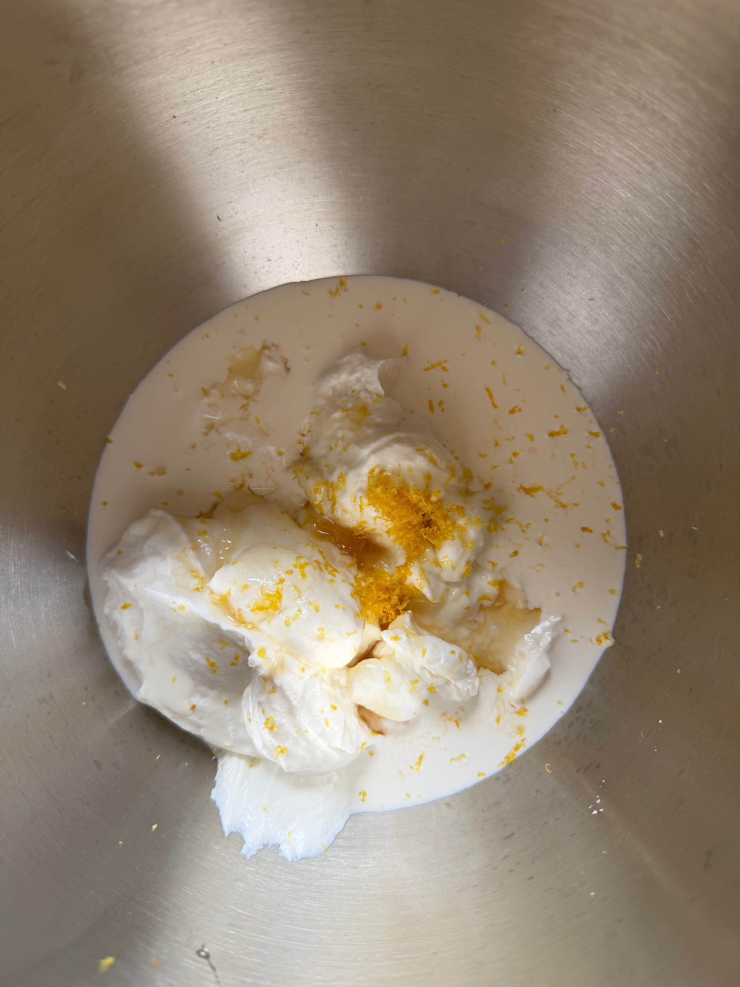 Cream, yogurt, and milk with grated lemon zest in a metal mixing bowl, ready to be mixed for a recipe.