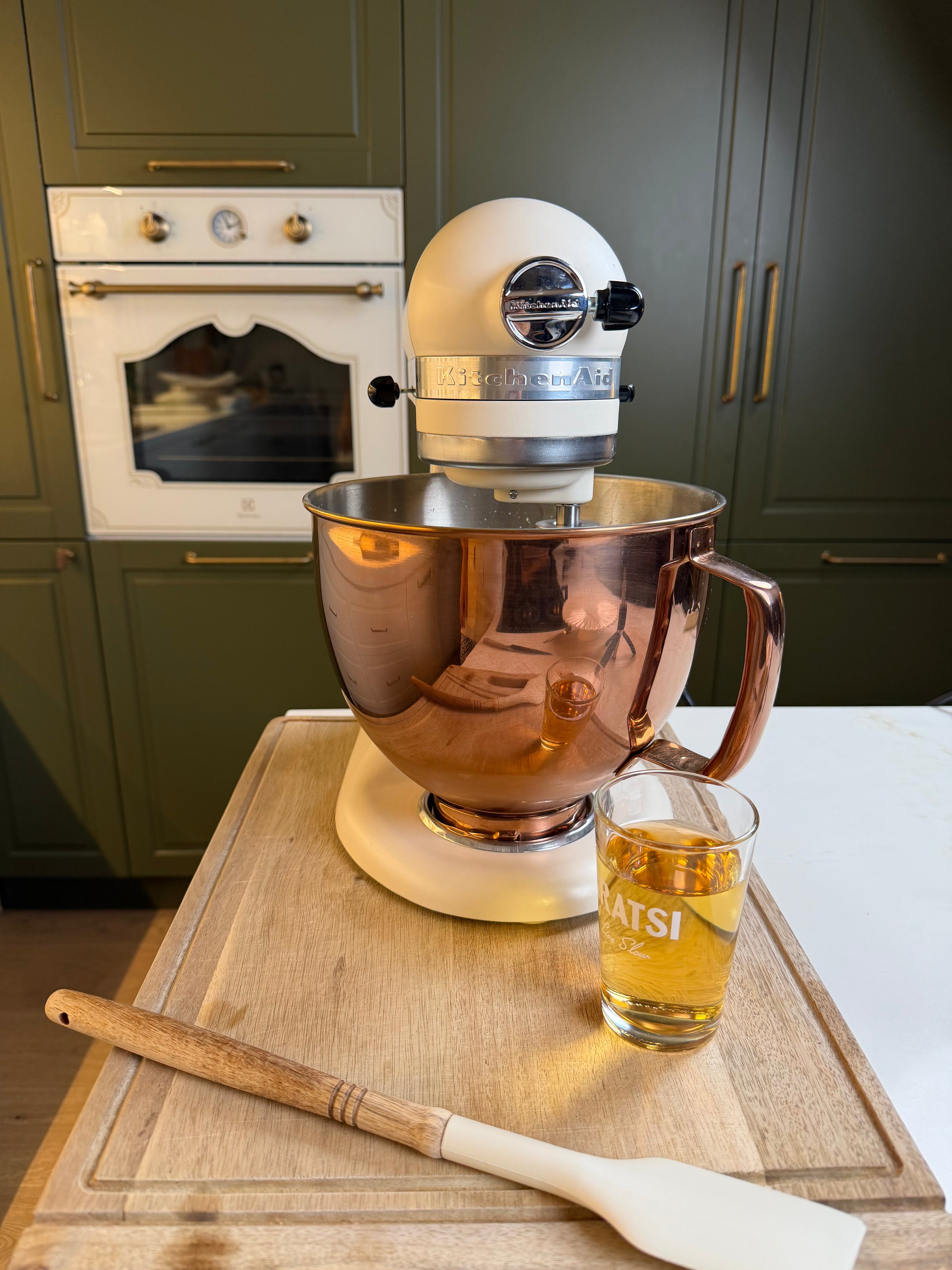 A stand mixer with a copper bowl sits on a wooden board. A spatula and a glass of amber liquid are beside it. Green cabinets and a white oven are in the background.