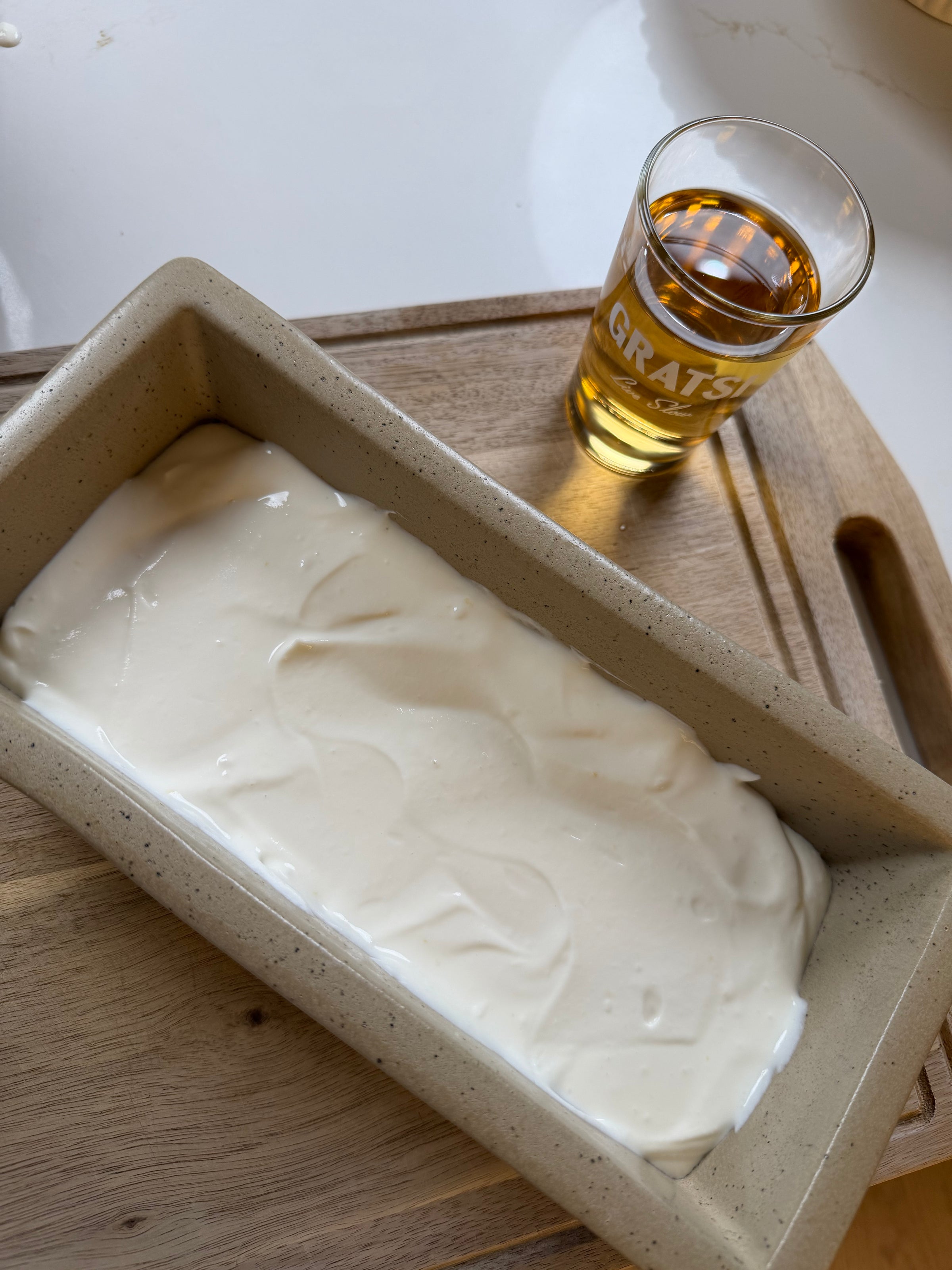 A rectangular baking dish with a creamy white mixture sits on a wooden cutting board next to a glass filled with a golden-brown liquid.