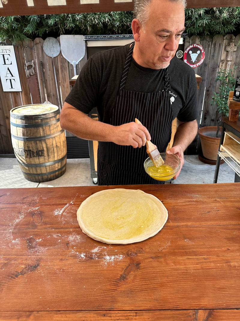 A man wearing a black apron brushes egg wash onto a round pizza dough on a wooden table in an outdoor kitchen, with a barrel and rustic decor in the background.