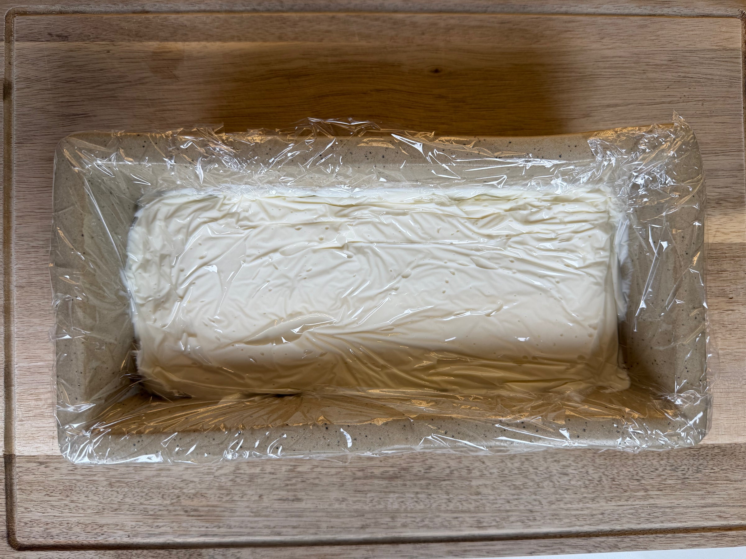 A rectangular block of cream cheese wrapped in plastic wrap sits in a loaf pan on a wooden surface.