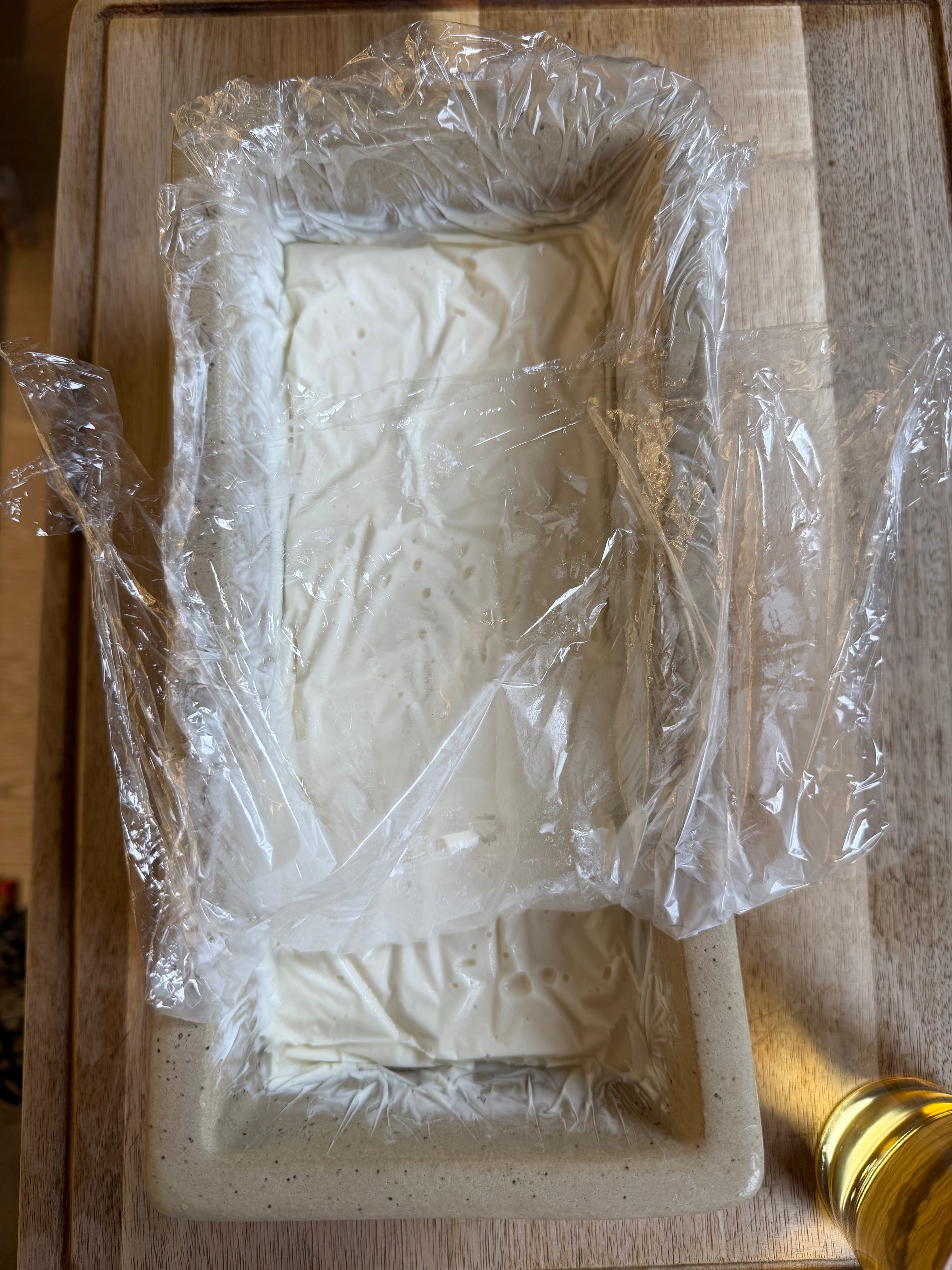 A rectangular dish lined with plastic wrap contains a block of smooth, white substance, possibly homemade ice cream or tofu. The wrap is partially peeled back, and the dish sits on a wooden surface. A yellow object is nearby.