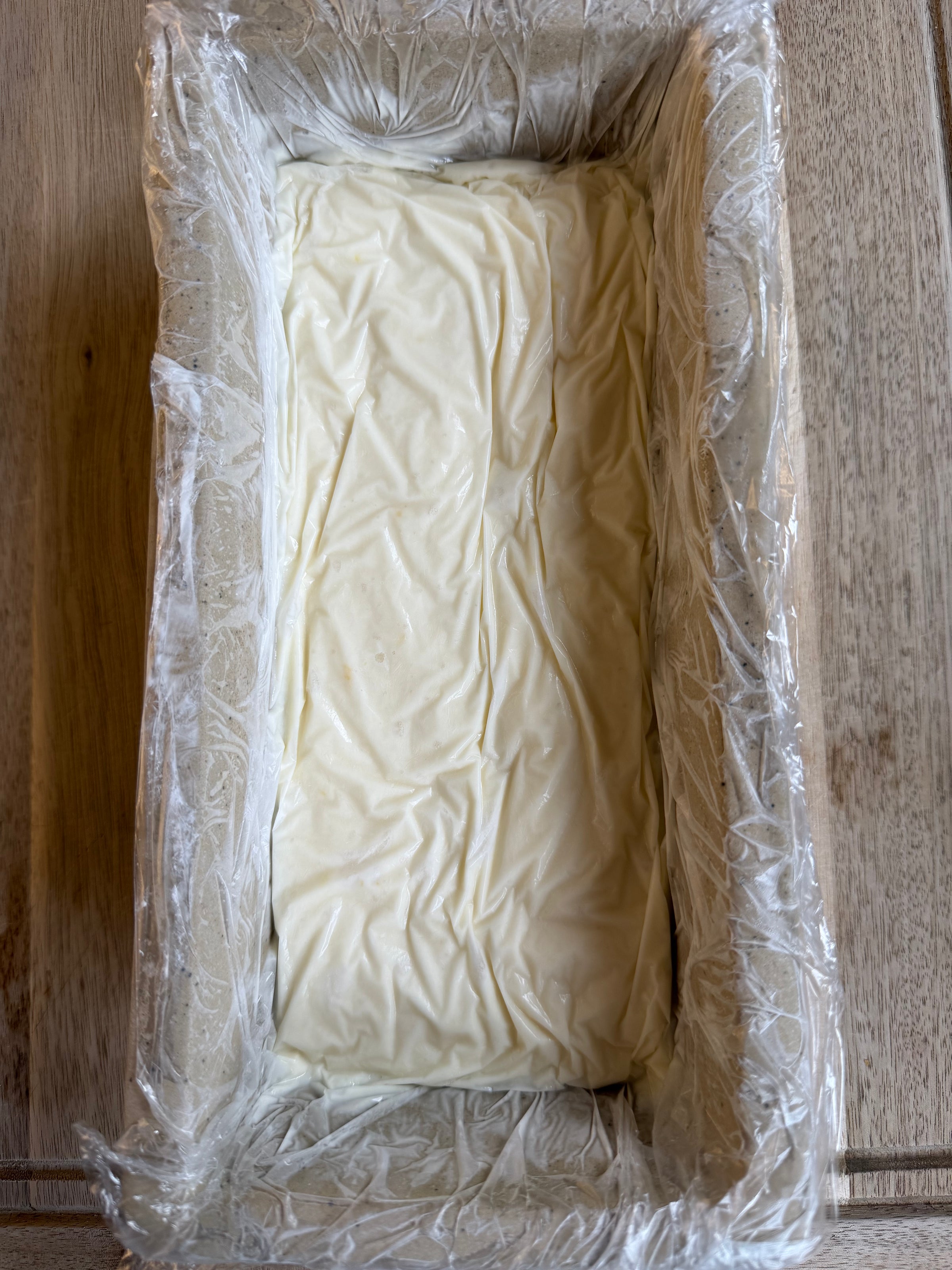 A rectangular loaf pan lined with plastic wrap, filled with a smooth, creamy white mixture, sitting on a wooden surface.