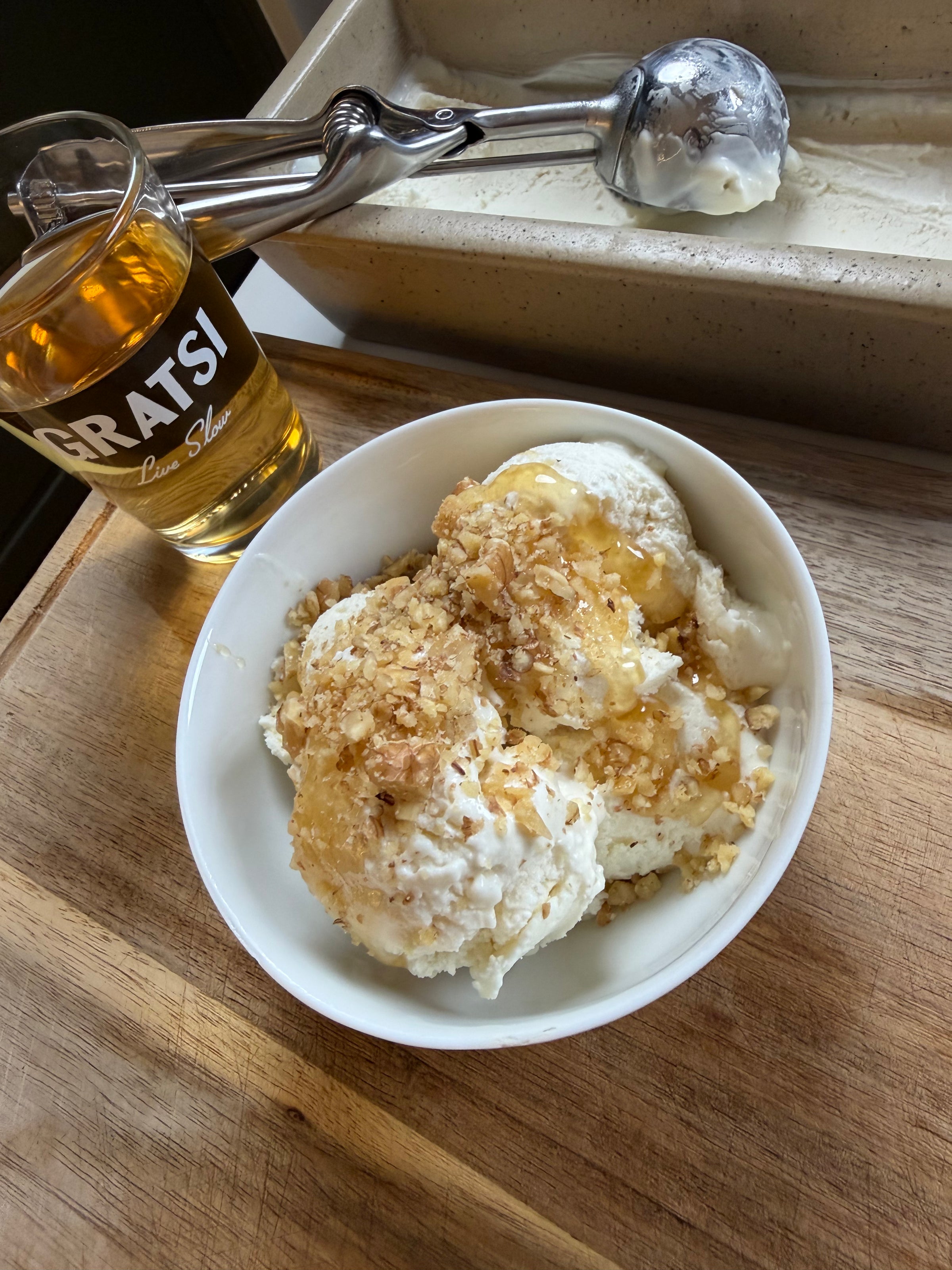 A bowl of vanilla ice cream topped with honey and chopped nuts sits on a wooden board. Next to it is a glass of amber liquid labeled GRATSI and a metal ice cream scoop rests in a container of ice cream.
