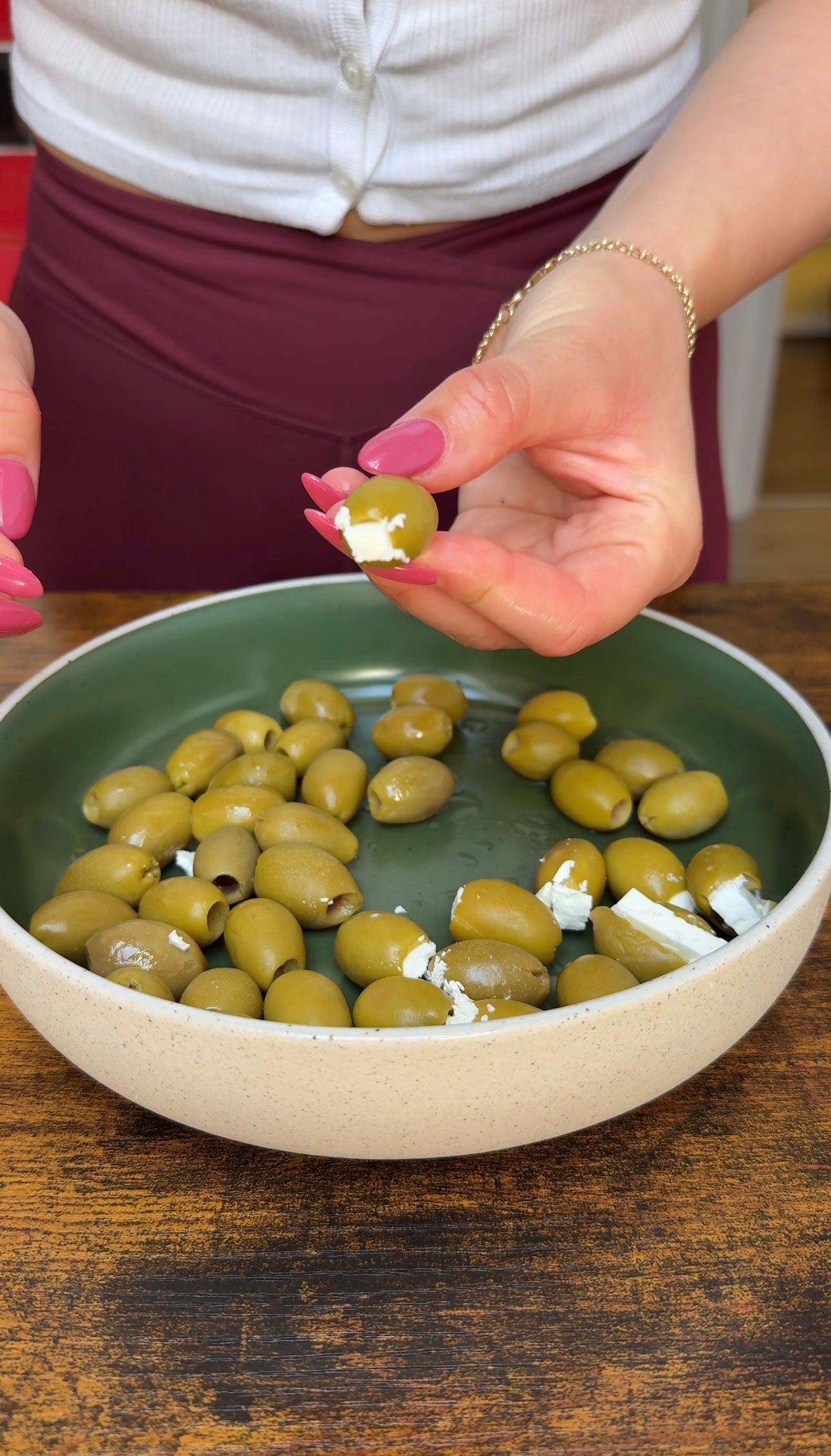 A person with pink nails holds a green olive stuffed with white cheese over a bowl filled with more stuffed green olives on a wooden table.