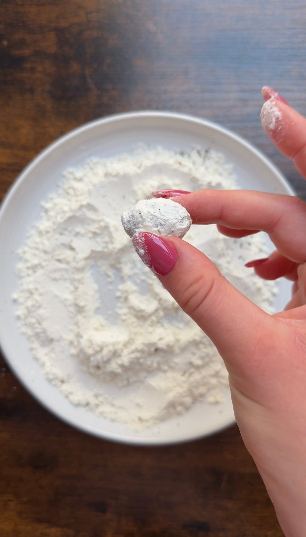 A hand with pink painted nails holds a small dough ball coated in flour above a white plate filled with flour on a wooden surface.
