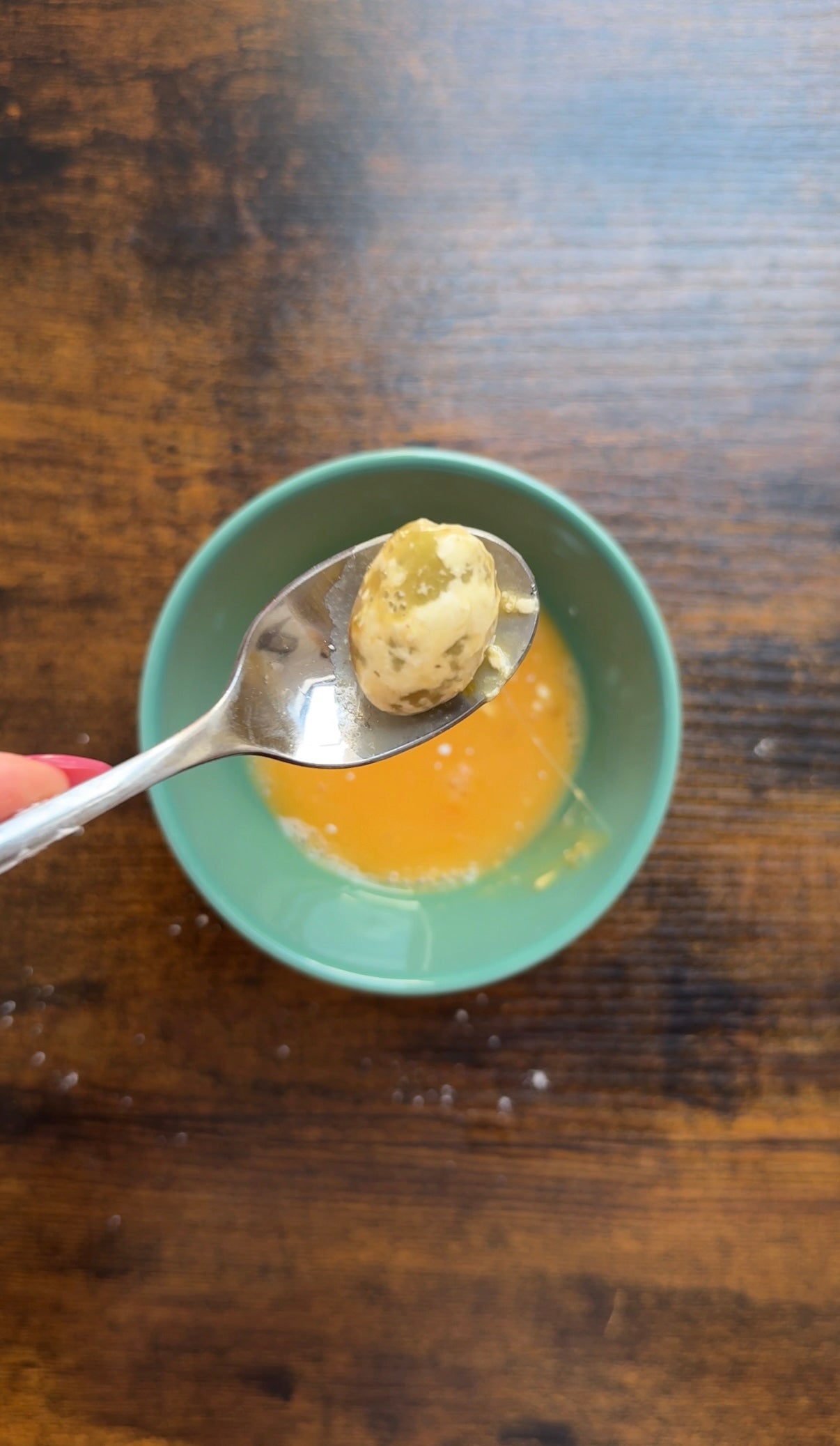 A spoon holding a small, marinated mozzarella ball is positioned over a teal bowl containing orange liquid, all set on a wooden table.