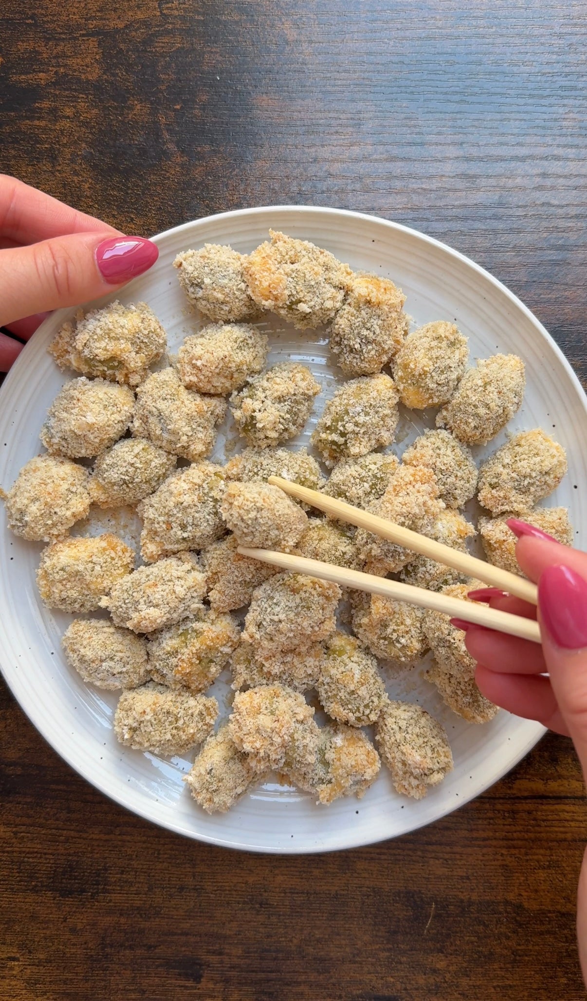 A plate of breaded, bite-sized snacks being held with chopsticks, with hands visible and nails painted pink on a wooden table.