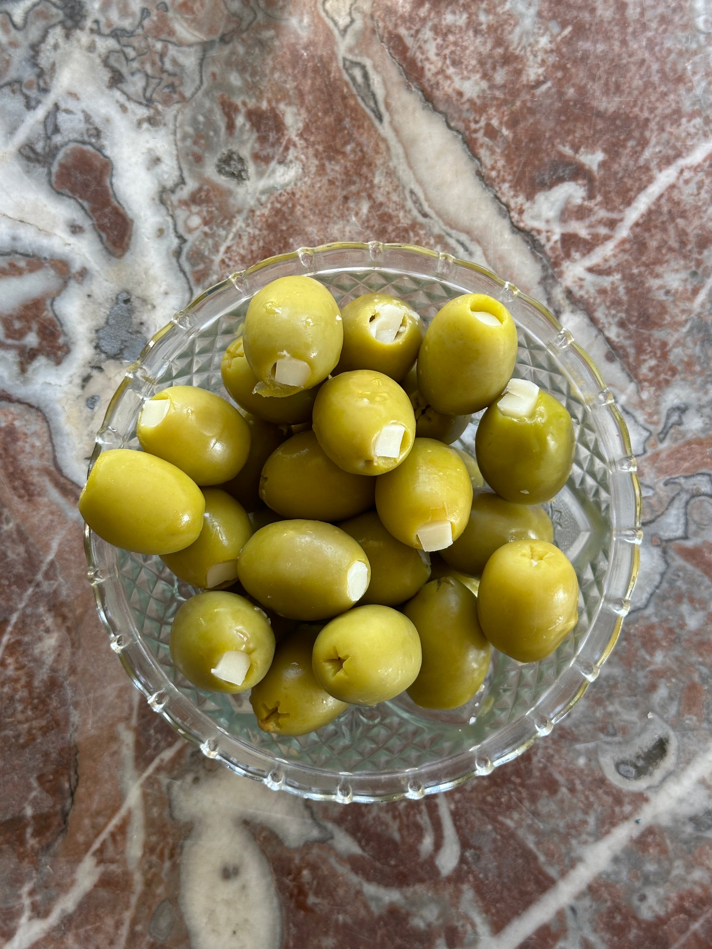 A glass bowl filled with green olives, each stuffed with a small piece of garlic, sits on a marbled surface.