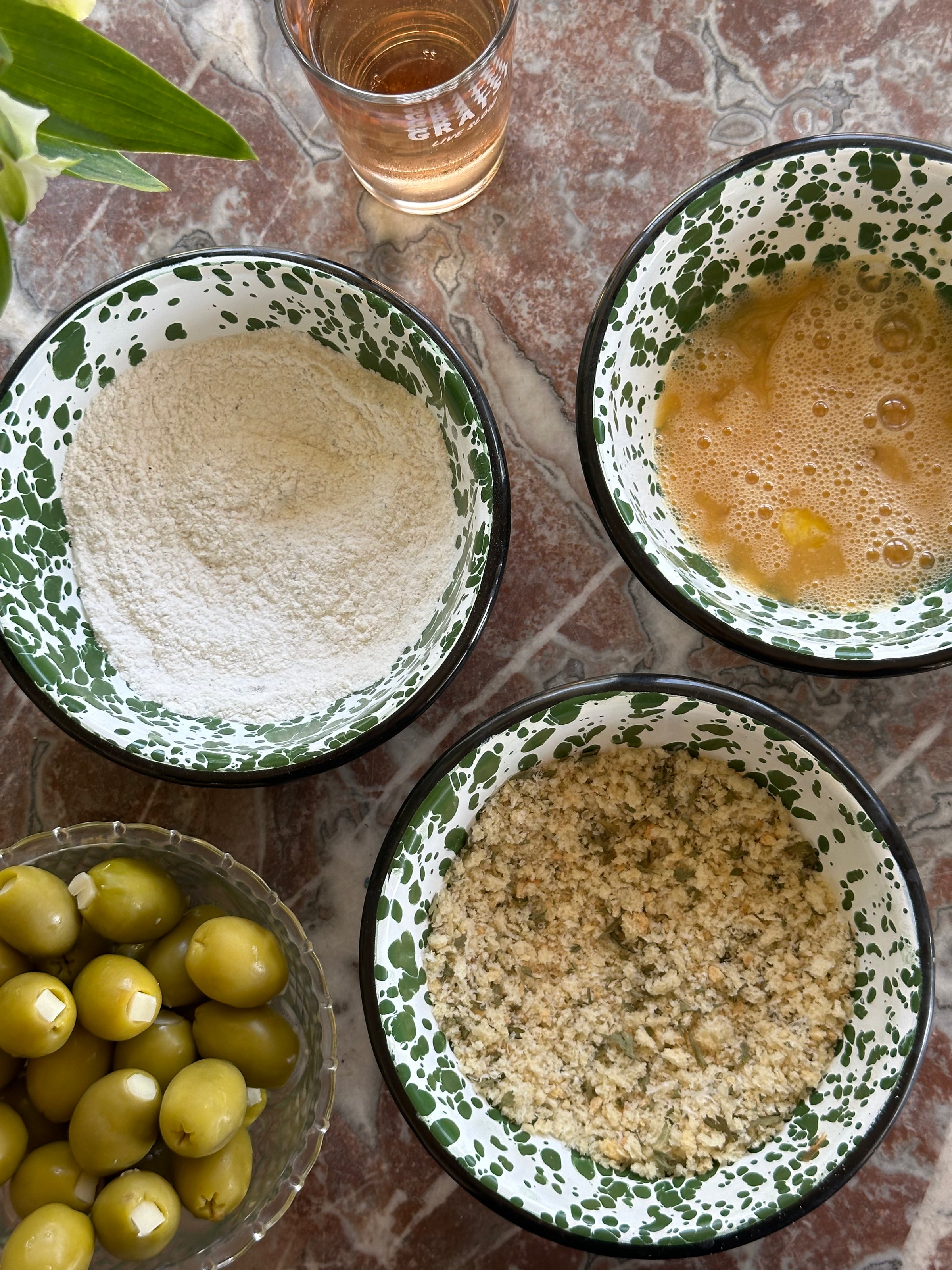 Three bowls on a marble surface: one with flour, one with beaten eggs, and one with seasoned breadcrumbs. A glass of pink liquid and a bowl of green olives are nearby. Green leaves are partially visible in the corner.