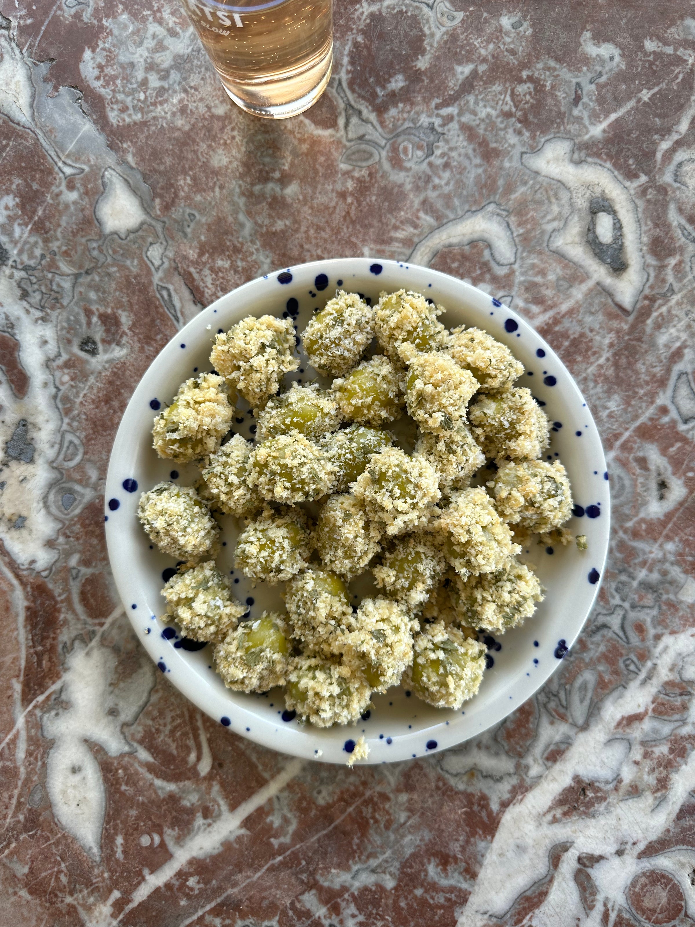 A white plate with blue speckles holds breaded green olives, set on a marble table. A glass with a light beverage is partially visible at the top of the image.