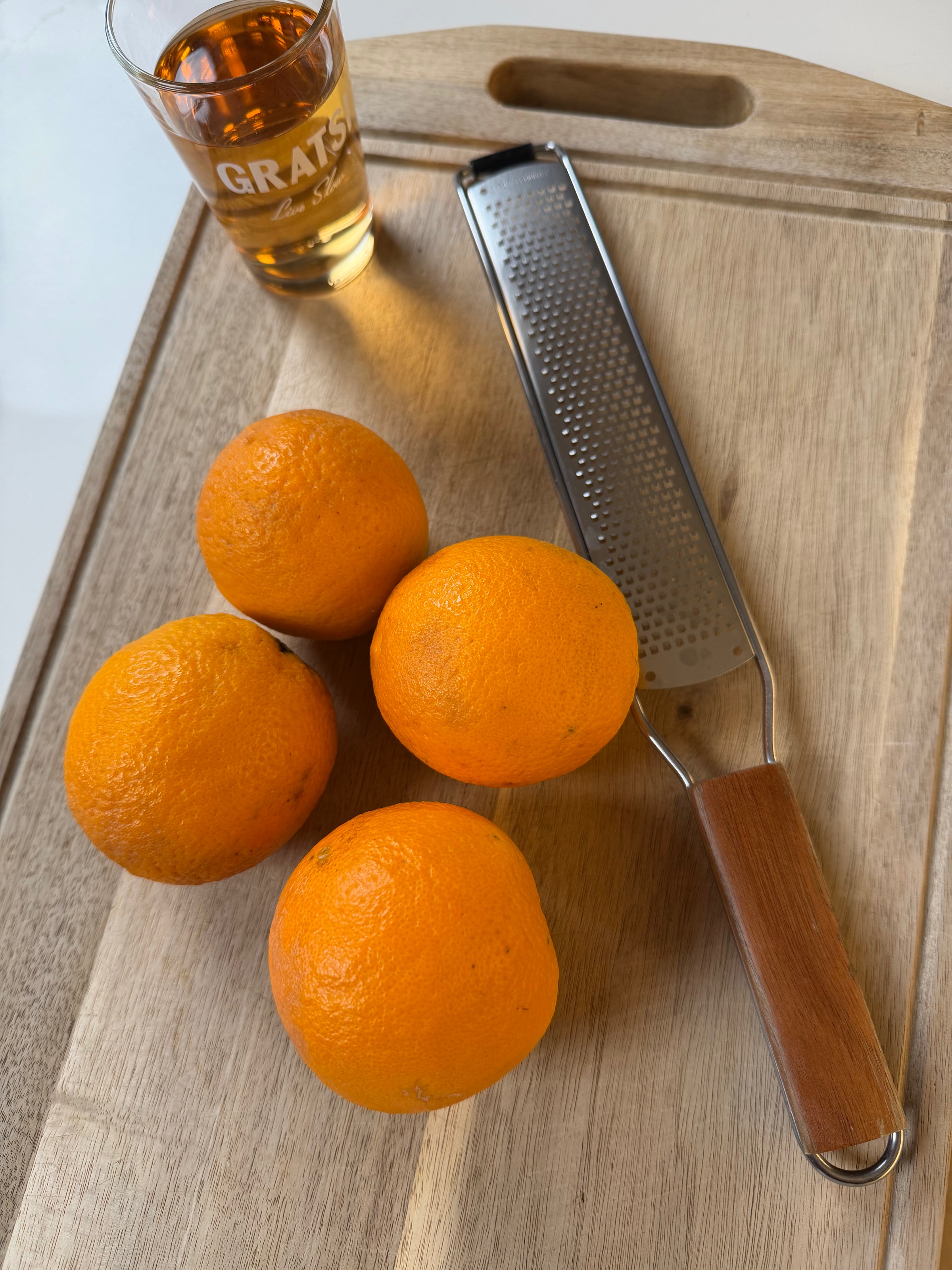 Four oranges and a metal zester with a wooden handle rest on a wooden cutting board. A glass of amber liquid with GRATS printed on it stands nearby.