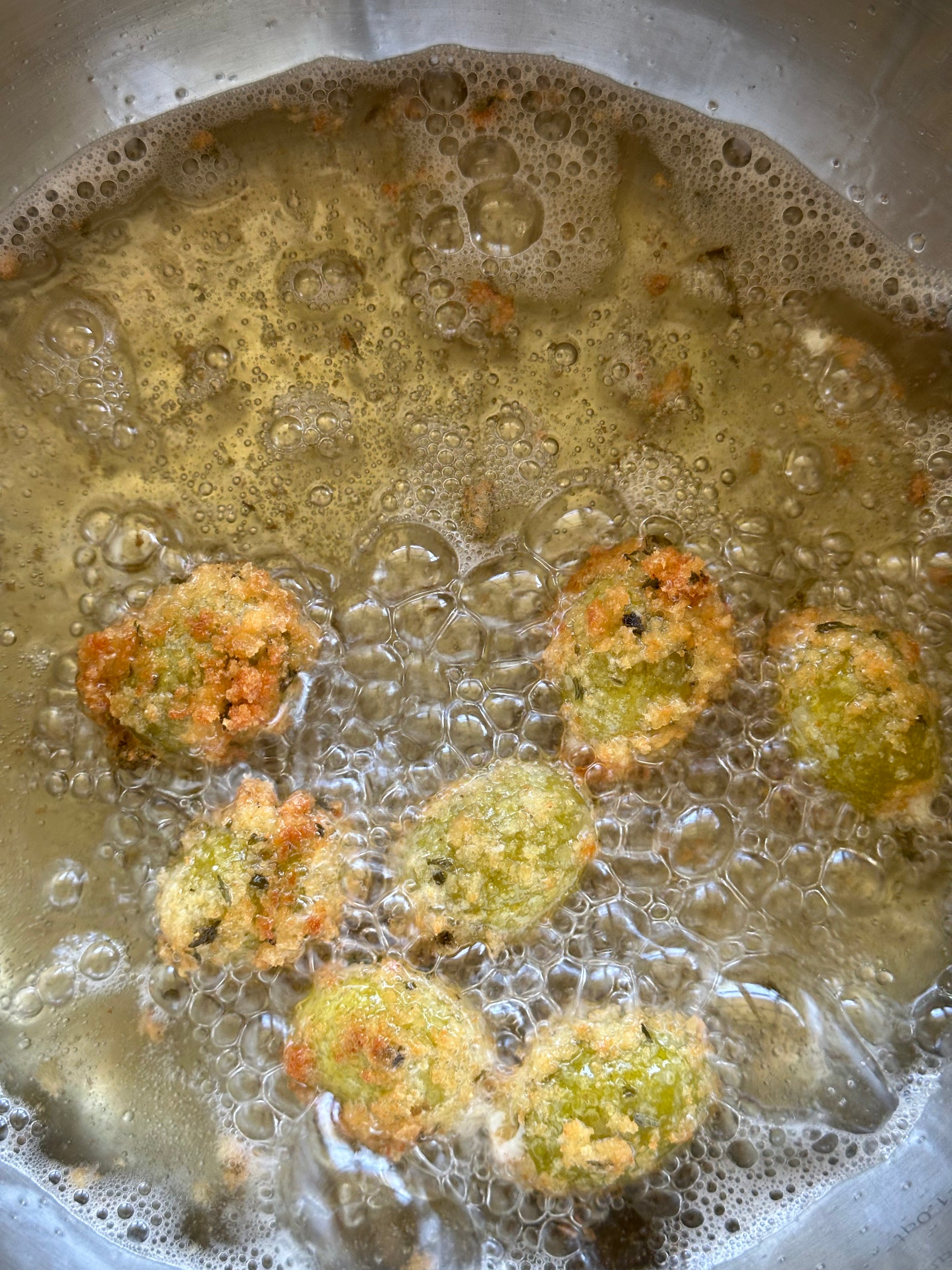 Seven green and golden fritters are frying in bubbling hot oil inside a metal pan. The oil is foaming slightly around the fritters as they cook.