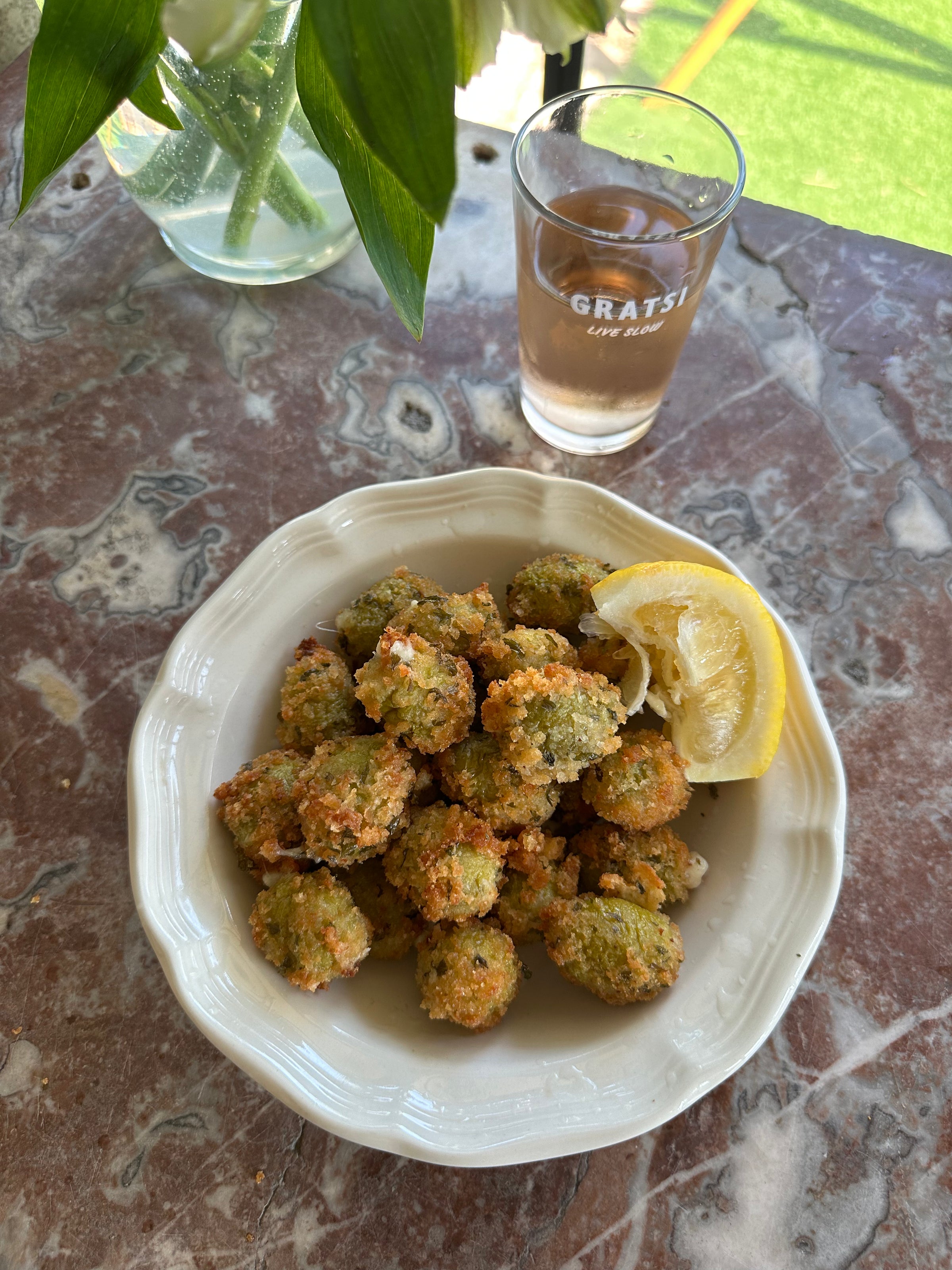 A white bowl filled with fried, breaded olives and a lemon wedge sits on a marble table. Behind the bowl is a glass with a brownish beverage, labeled GRATSI. Greenery is visible in the background.