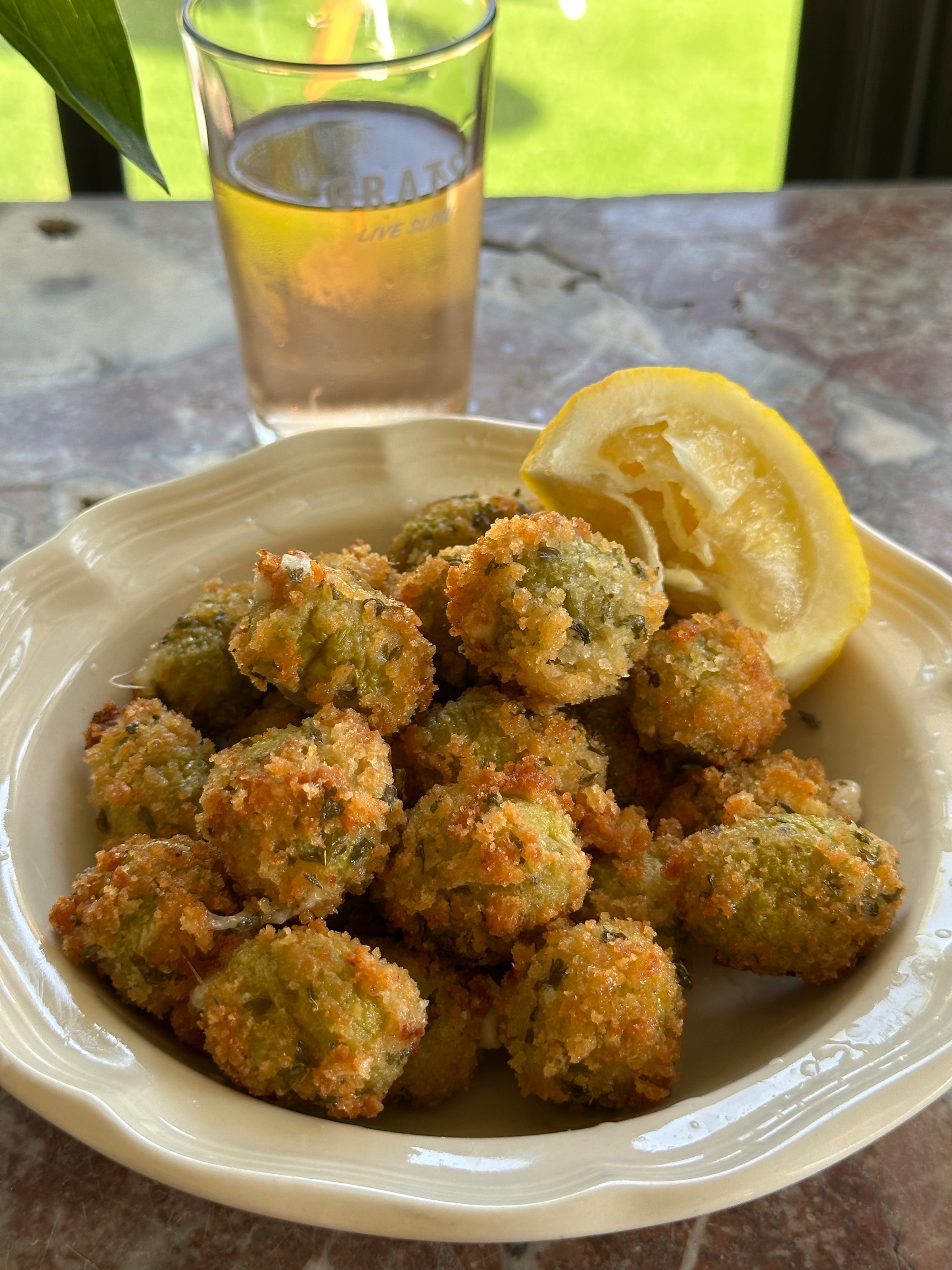 A bowl of golden-brown fried balls garnished with a lemon wedge, served on a white plate. A glass of light-colored drink sits in the background on a marble table by a sunny window.