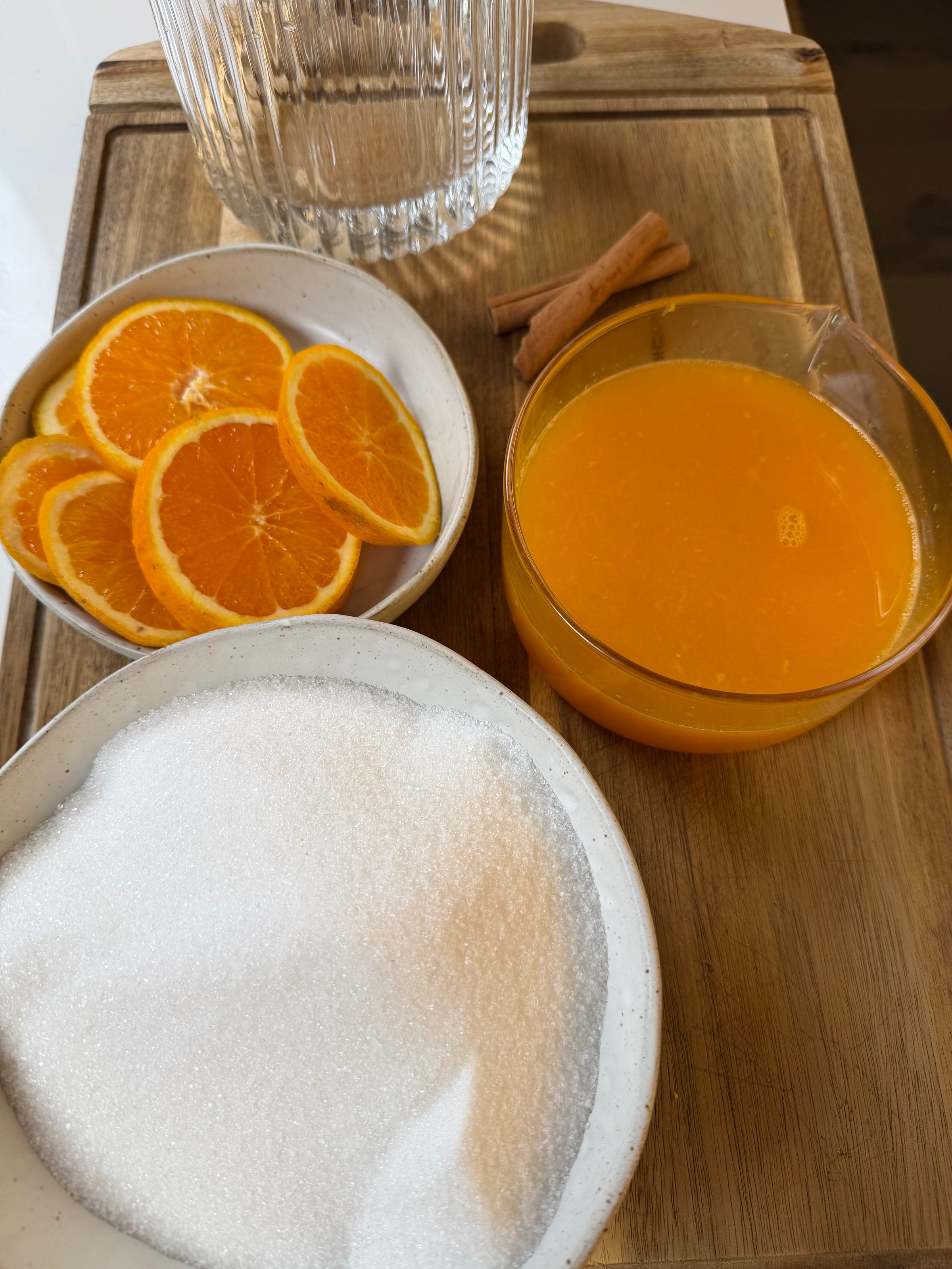 A wooden tray with a bowl of sliced oranges, a bowl of white sugar, a glass of orange juice, two cinnamon sticks, and a glass pitcher.