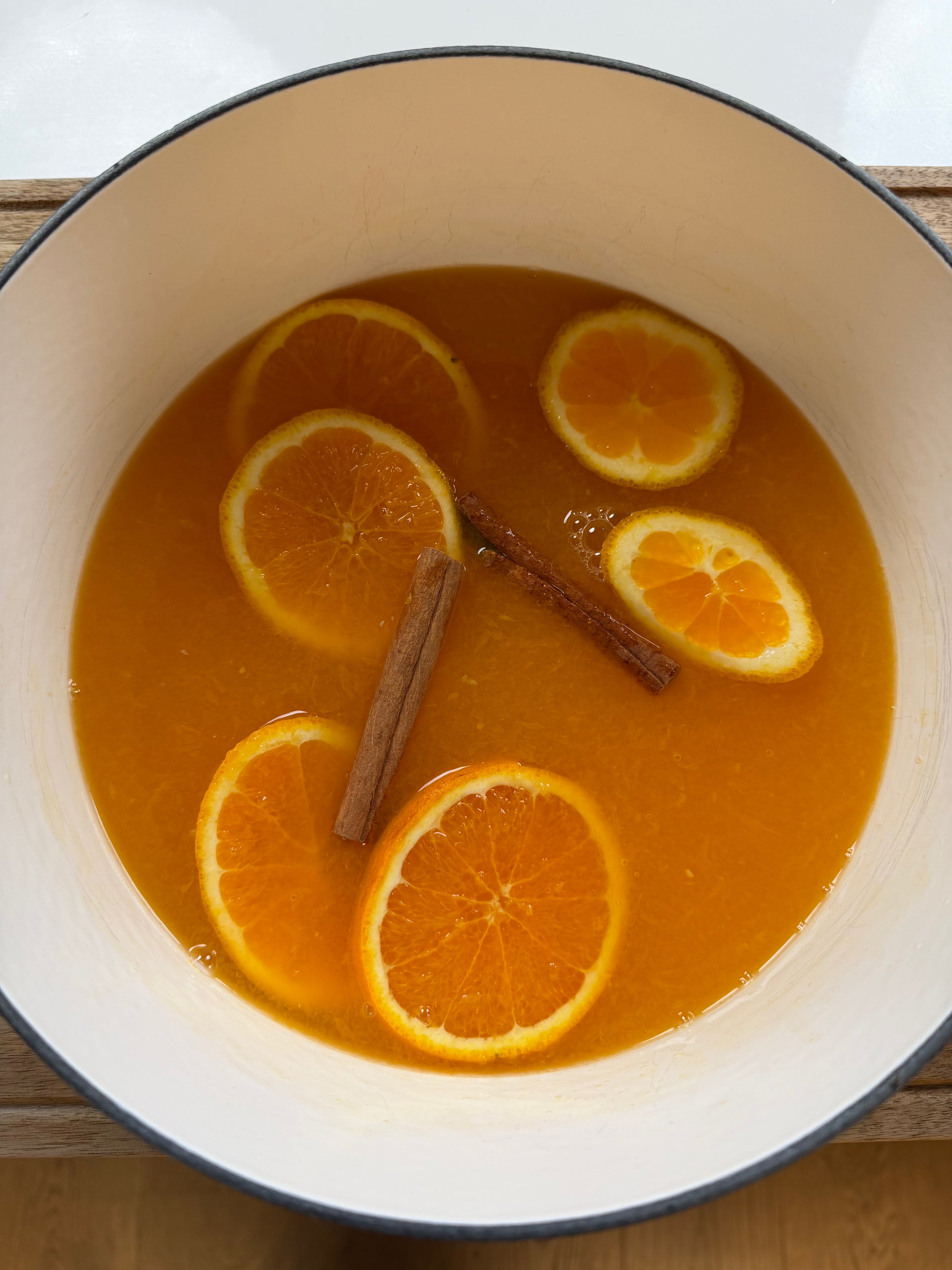 A white pot filled with orange liquid, orange slices, and two cinnamon sticks floating on top.