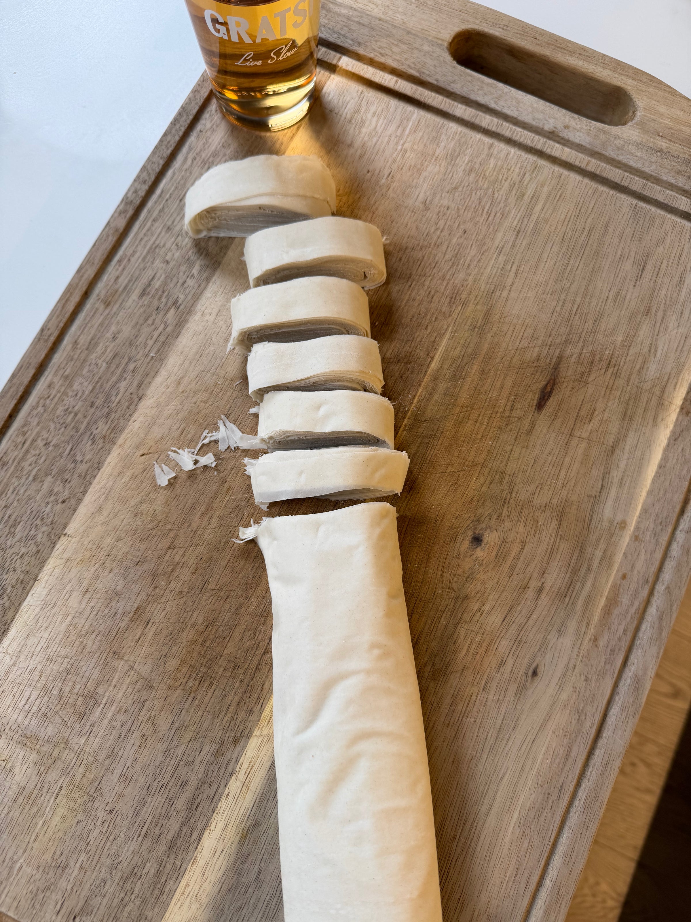 Sliced dough roll on a wooden cutting board, with a glass containing a light brown drink in the background.