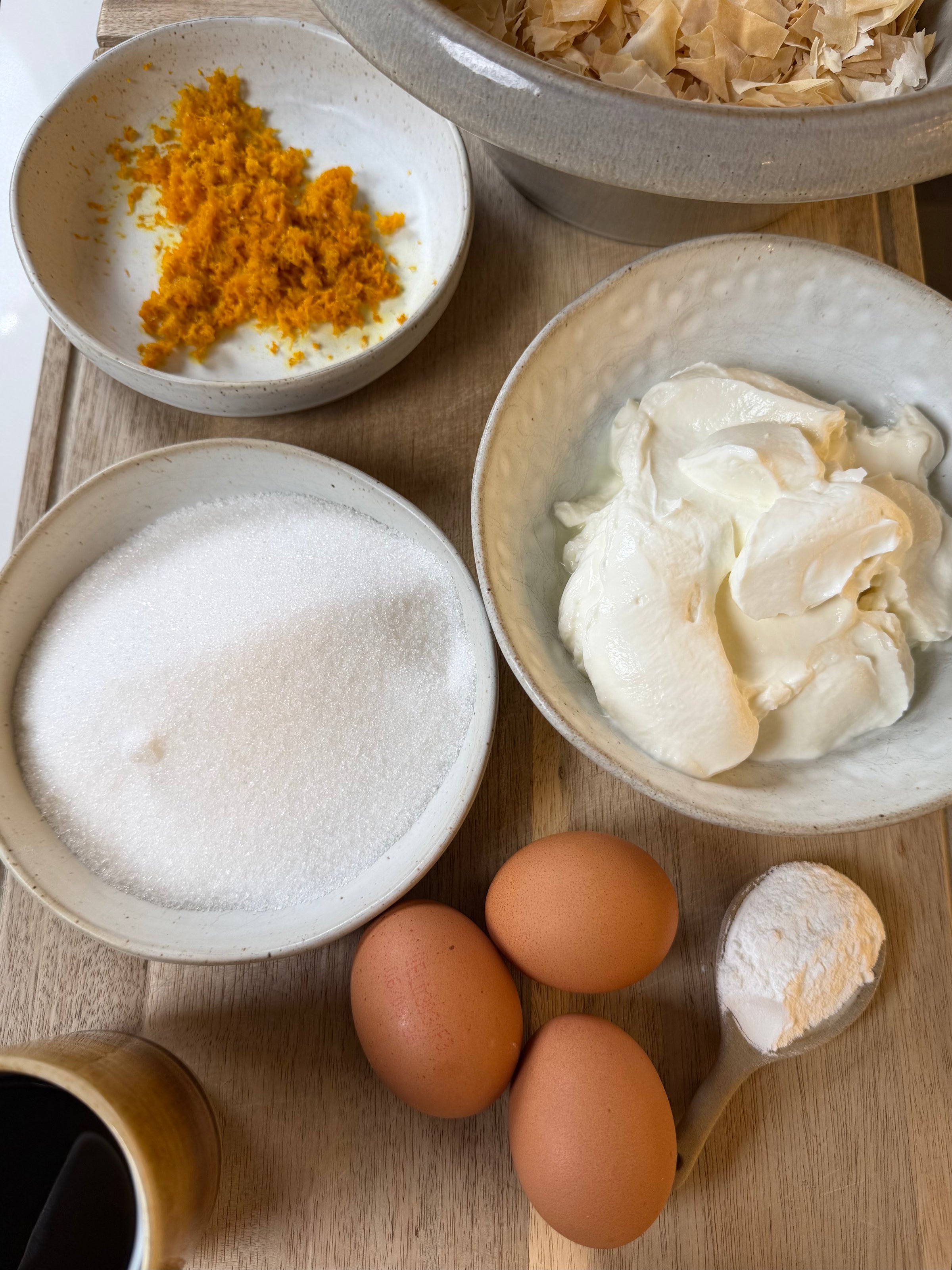 Three brown eggs, a bowl of granulated sugar, a dish of orange zest, a bowl of whipped cream cheese, a spoonful of flour, and a small cup of dark liquid are arranged on a wooden surface.