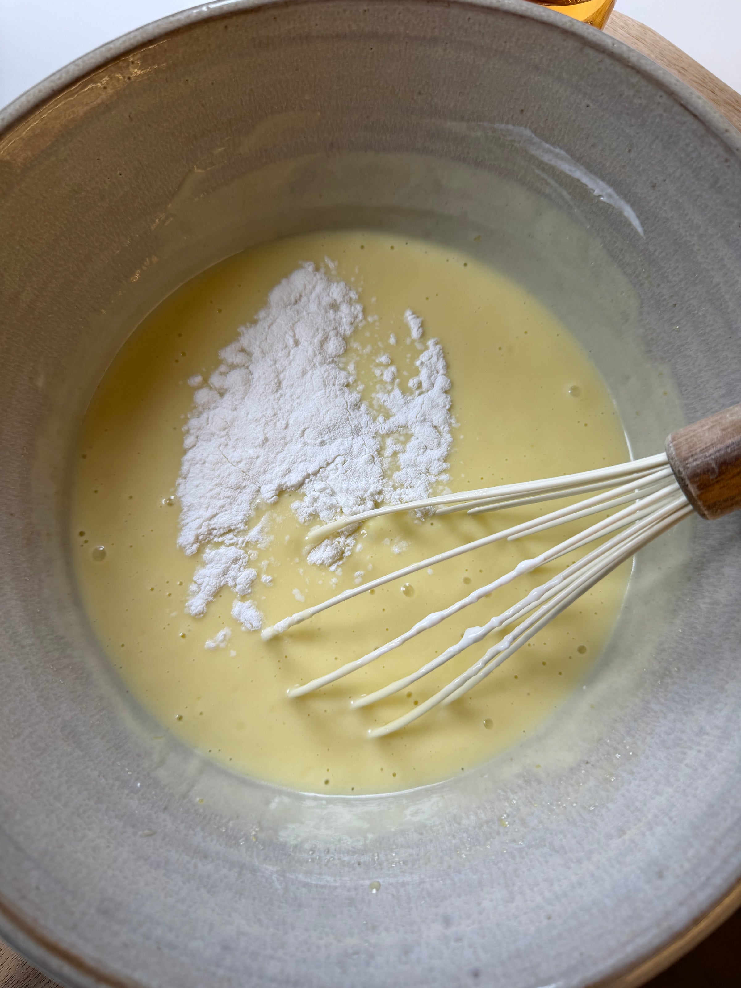 A metal whisk rests in a bowl of pale yellow batter with a mound of white flour or powdered sugar on top, ready to be mixed in.
