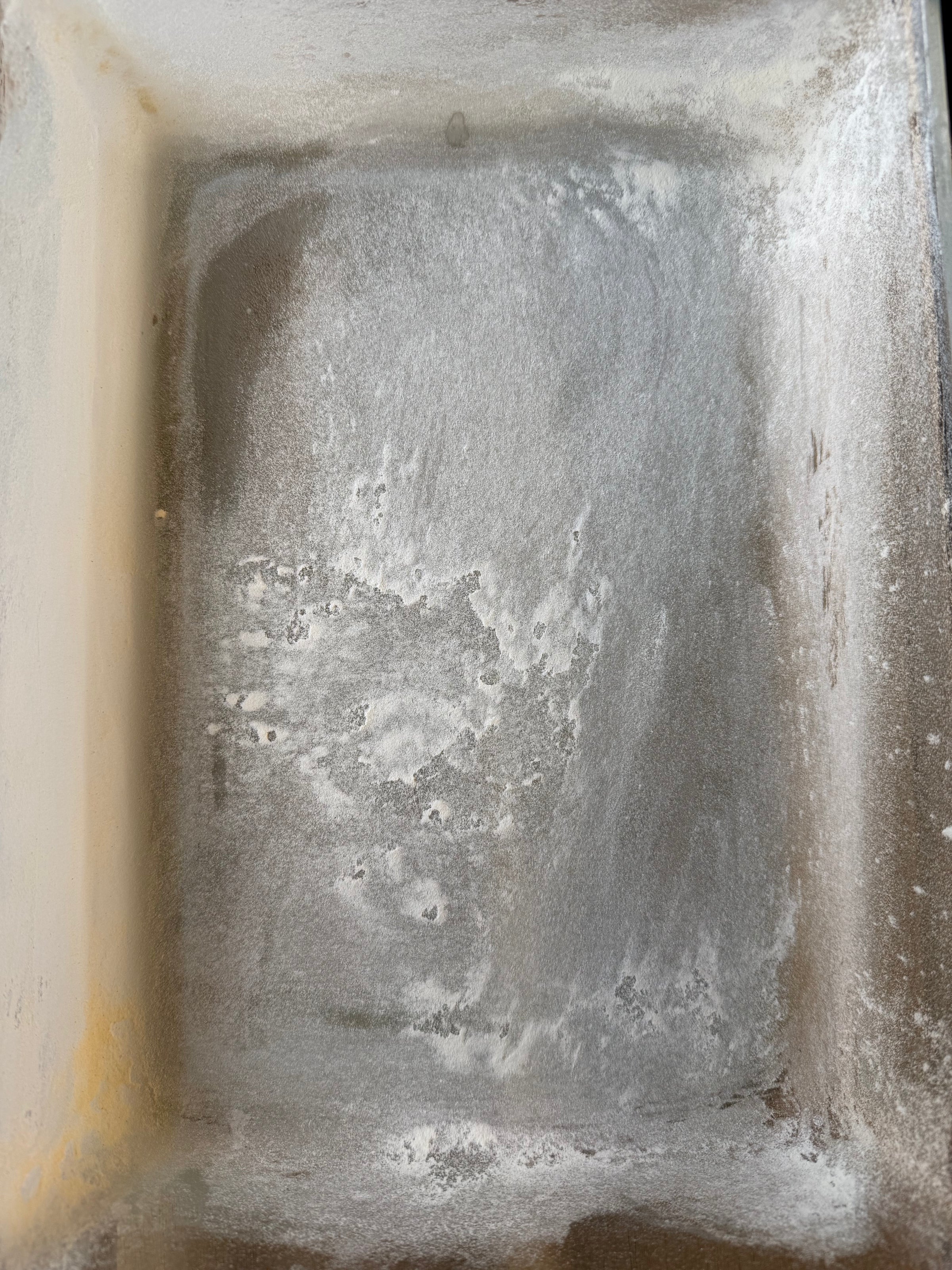 A rectangular metal baking pan with a light dusting of flour on its surface, likely prepared for baking. The edges and corners of the pan are visible, and the flour is unevenly spread.