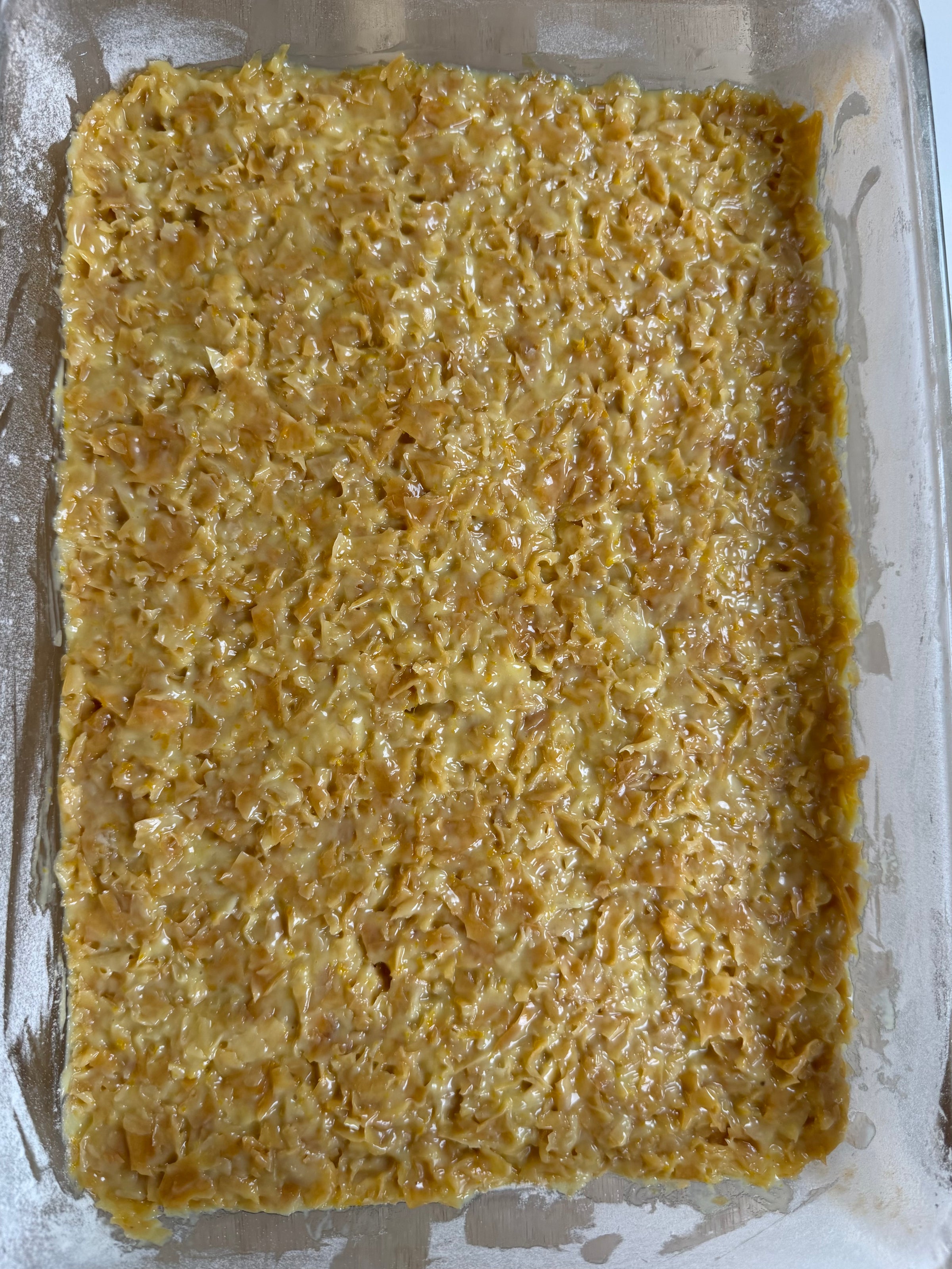 A glass baking dish filled with an uncooked, yellowish, crunchy cereal mixture spread evenly to the edges, likely prepared for baking cereal treats or bars.