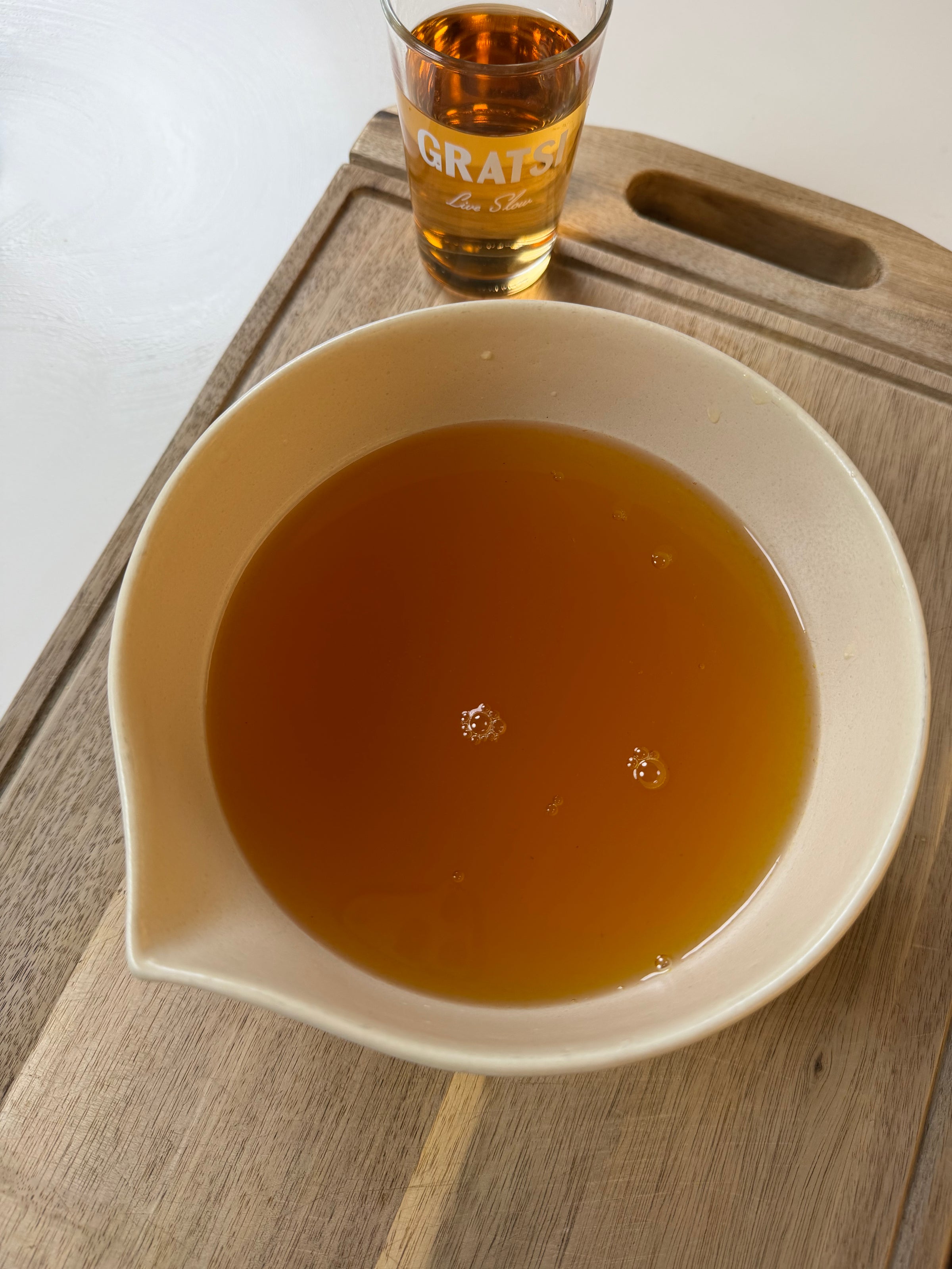 A white bowl filled with amber-colored liquid sits on a wooden tray. In the background, there is a small glass with similar liquid and the word GRATSI written on it.