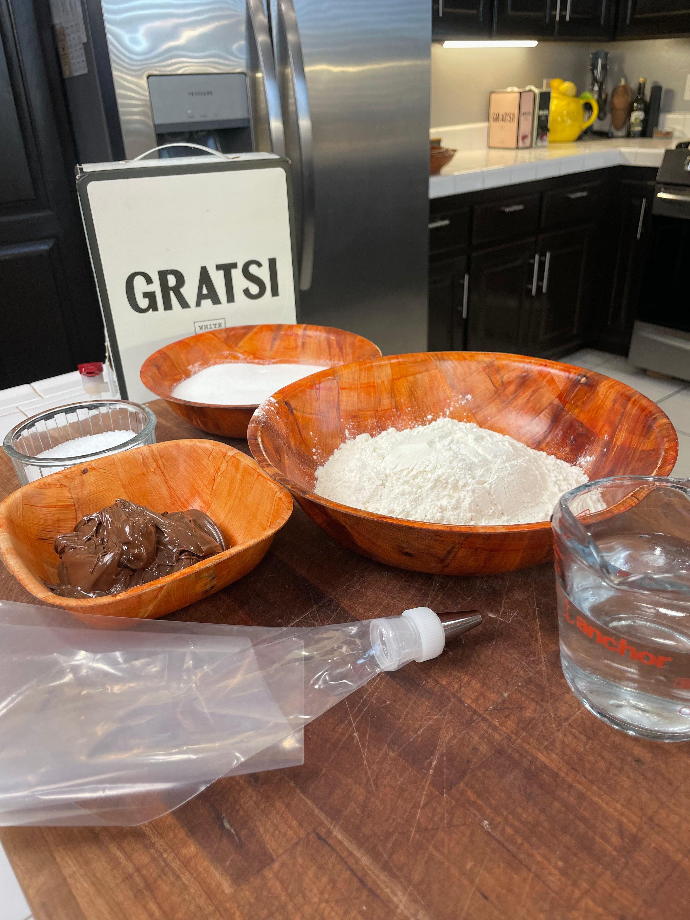 Various baking ingredients are arranged on a wooden counter, including flour, sugar, chocolate spread, water in a glass measuring cup, a piping bag with a tip, and a “GRATSI” sign in a modern kitchen.