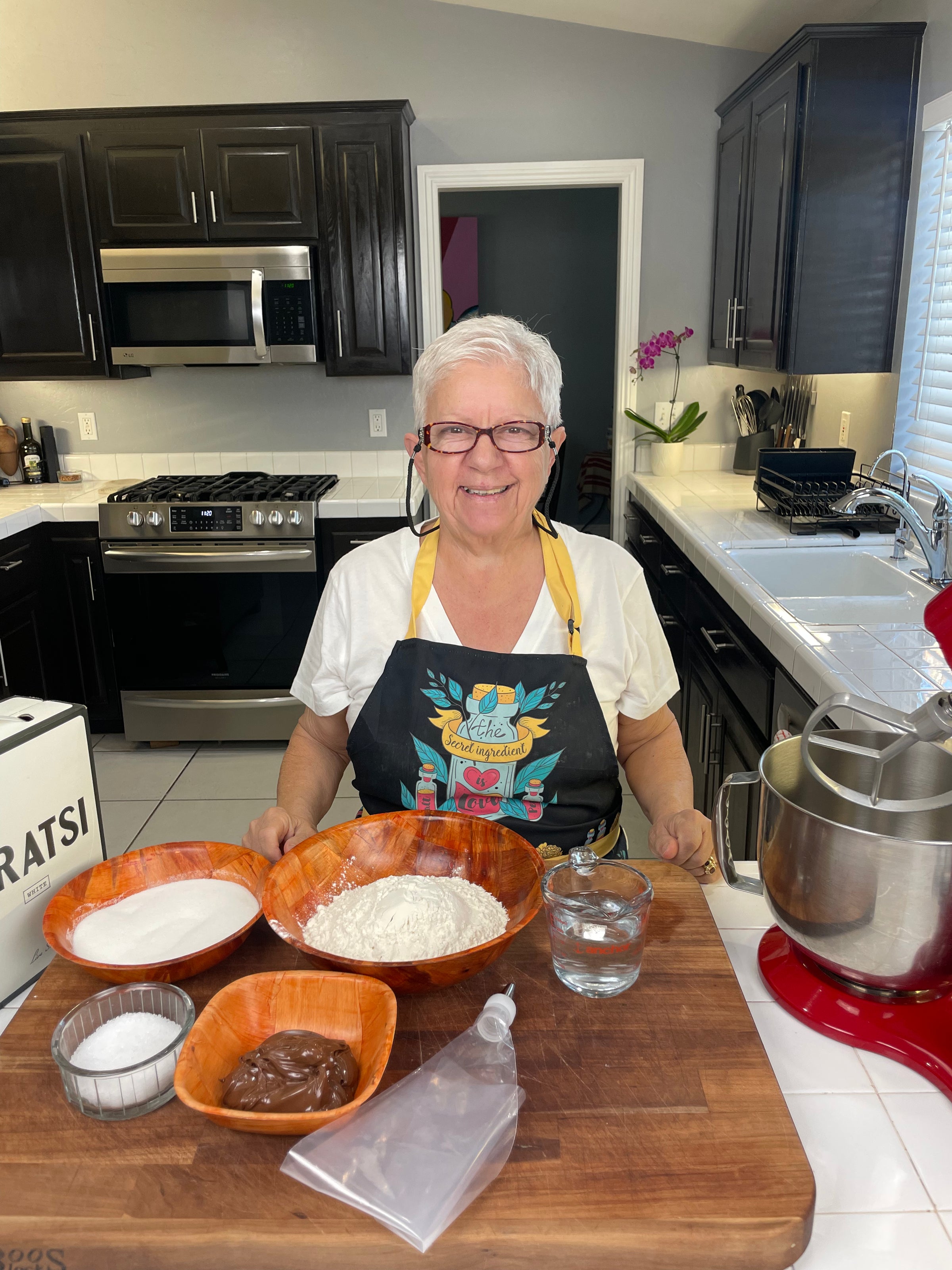 A smiling older woman with short white hair stands in a kitchen behind a counter with bowls of flour, sugar, water, chocolate spread, and a piping bag. She wears glasses, an apron, and a T-shirt. Kitchen appliances are visible in the background.