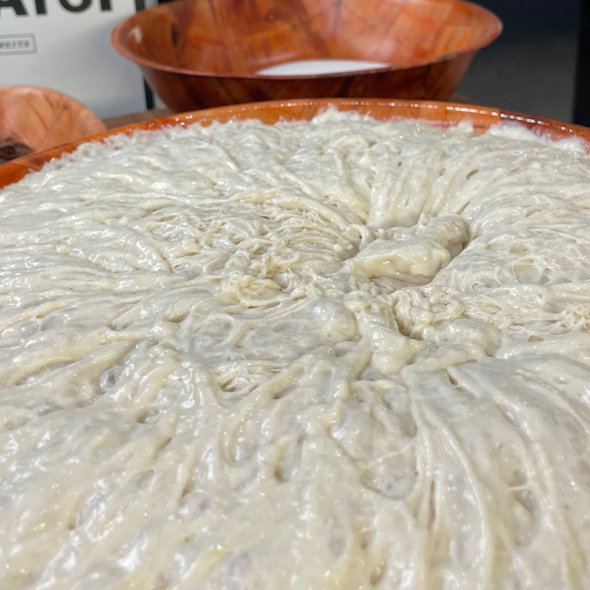 A close-up view of risen, bubbly bread dough in a large orange bowl, showing a stretchy, airy texture. In the background, additional bowls and a box are partially visible.