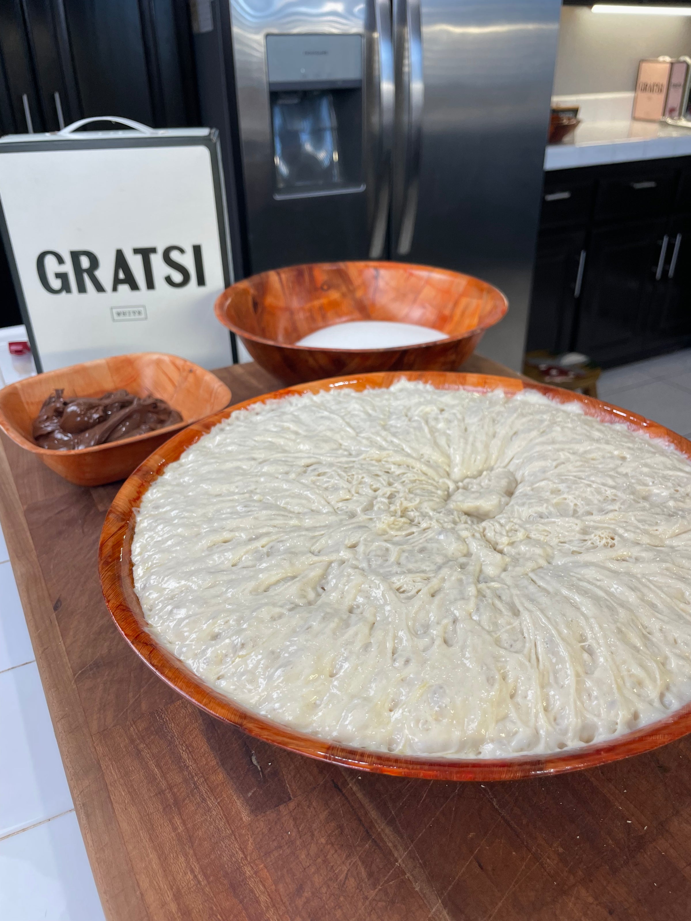 Three orange bowls on a kitchen counter: one with bubbly rising bread dough, one with chocolate spread, and one with white flour. A refrigerator and a sign that reads GRATSI are in the background.