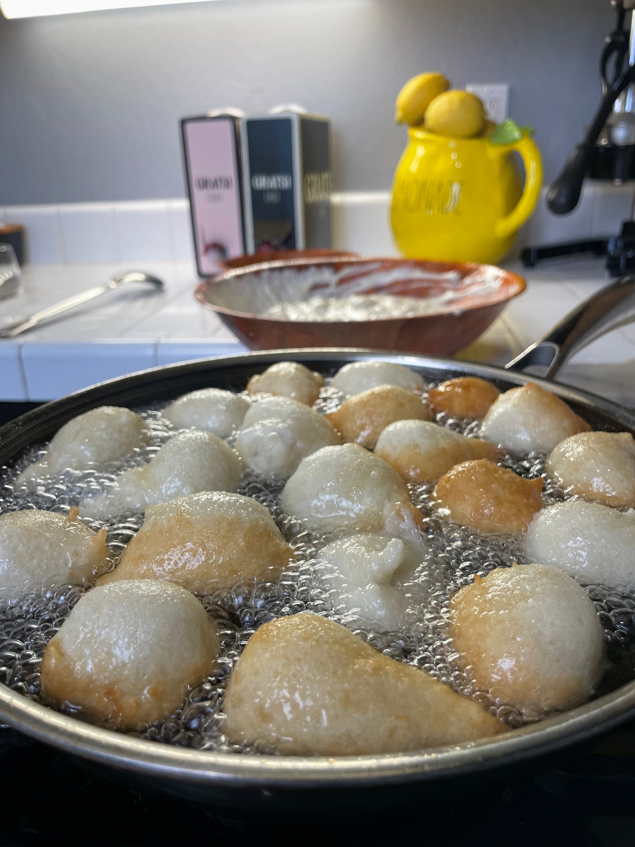 Round dough balls frying in hot oil in a pan on a stovetop, with a mixing bowl and a yellow lemon-themed container in the background on a kitchen counter.