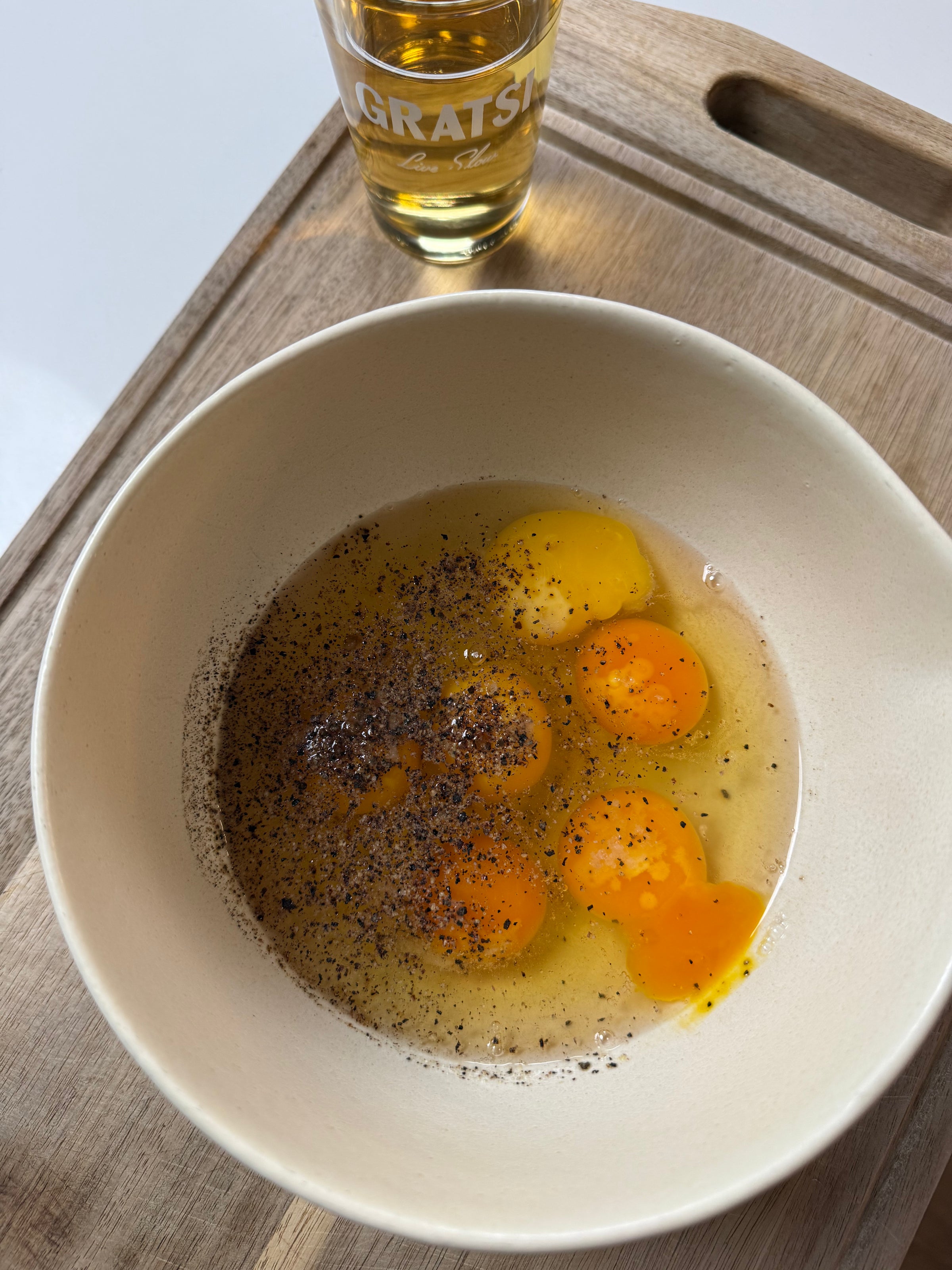 A white bowl with four cracked eggs and black pepper sits on a wooden tray. In the background, there is a glass containing a golden liquid.