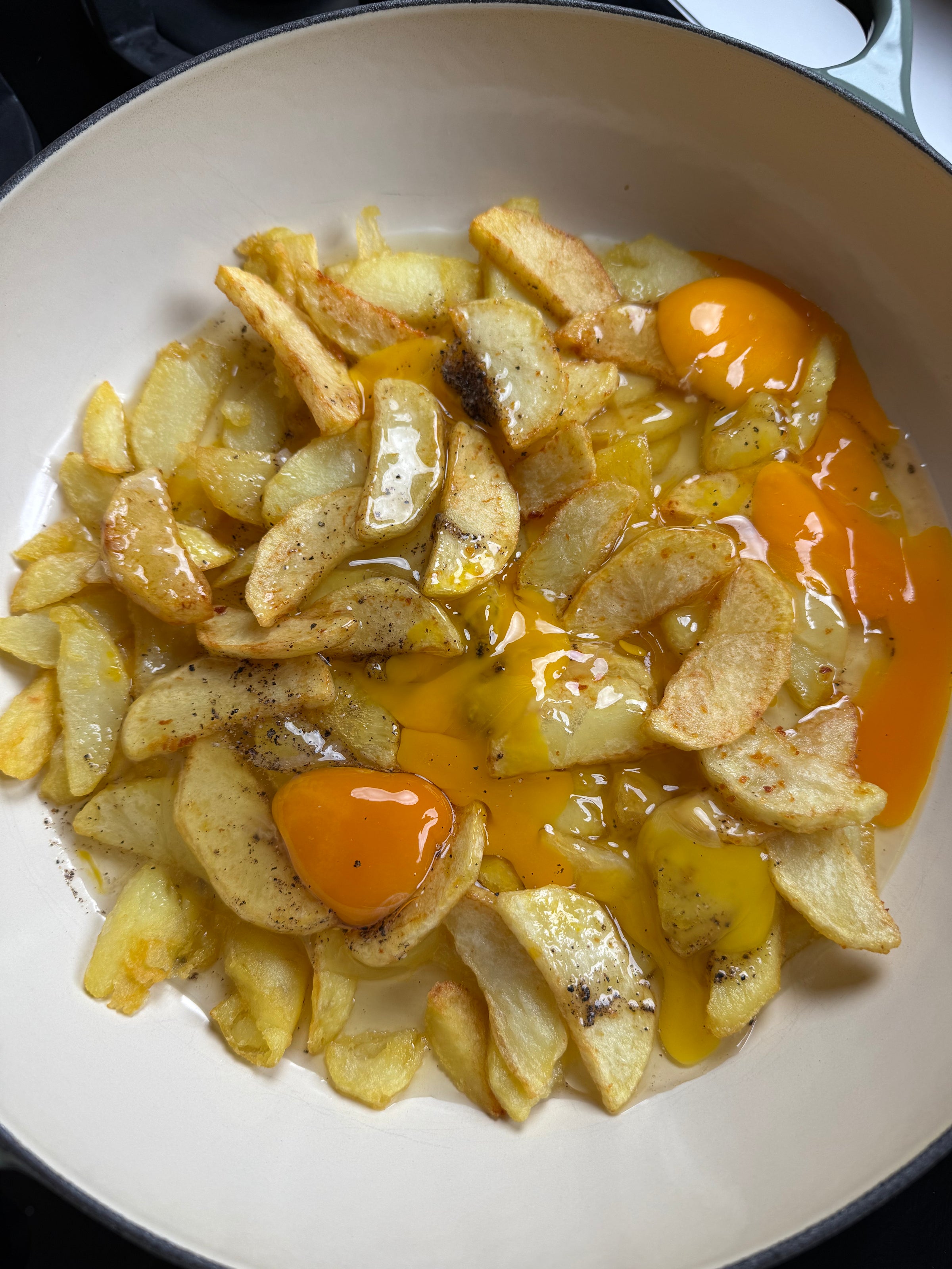 Sliced potatoes are frying in a pan with several cracked eggs on top. The egg yolks are partially broken, mixing slightly with the potatoes. The dish is lightly seasoned with salt and pepper.