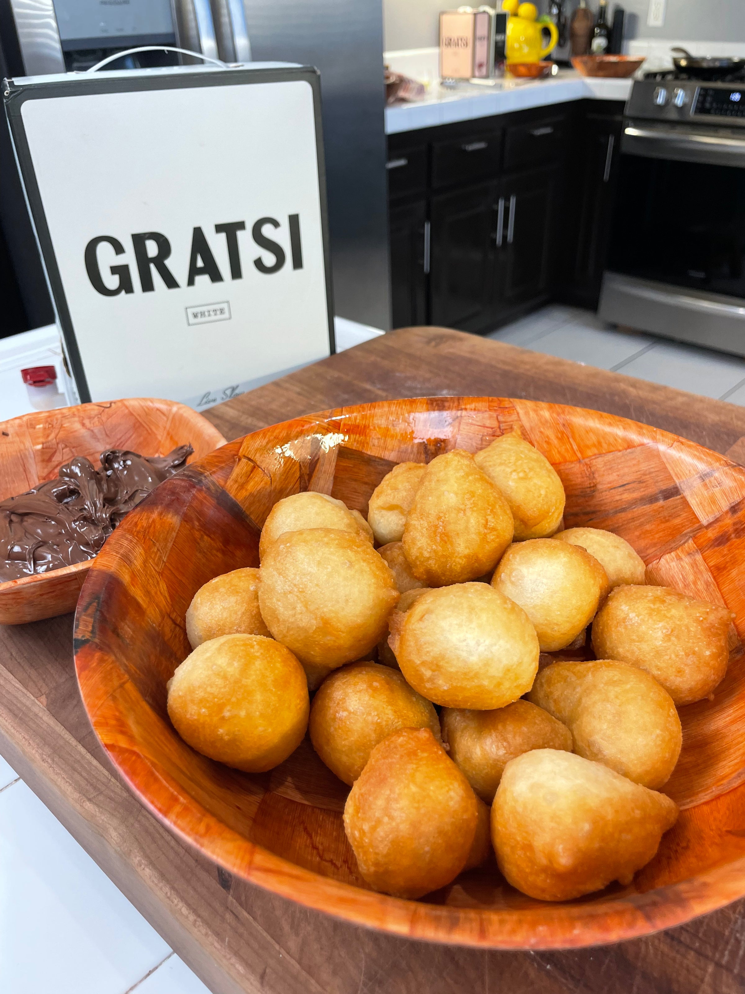 A wooden bowl filled with golden, round fried dough balls sits on a kitchen counter near a small dish of chocolate spread. In the background, a white GRATS! box is visible next to various kitchen items and appliances.