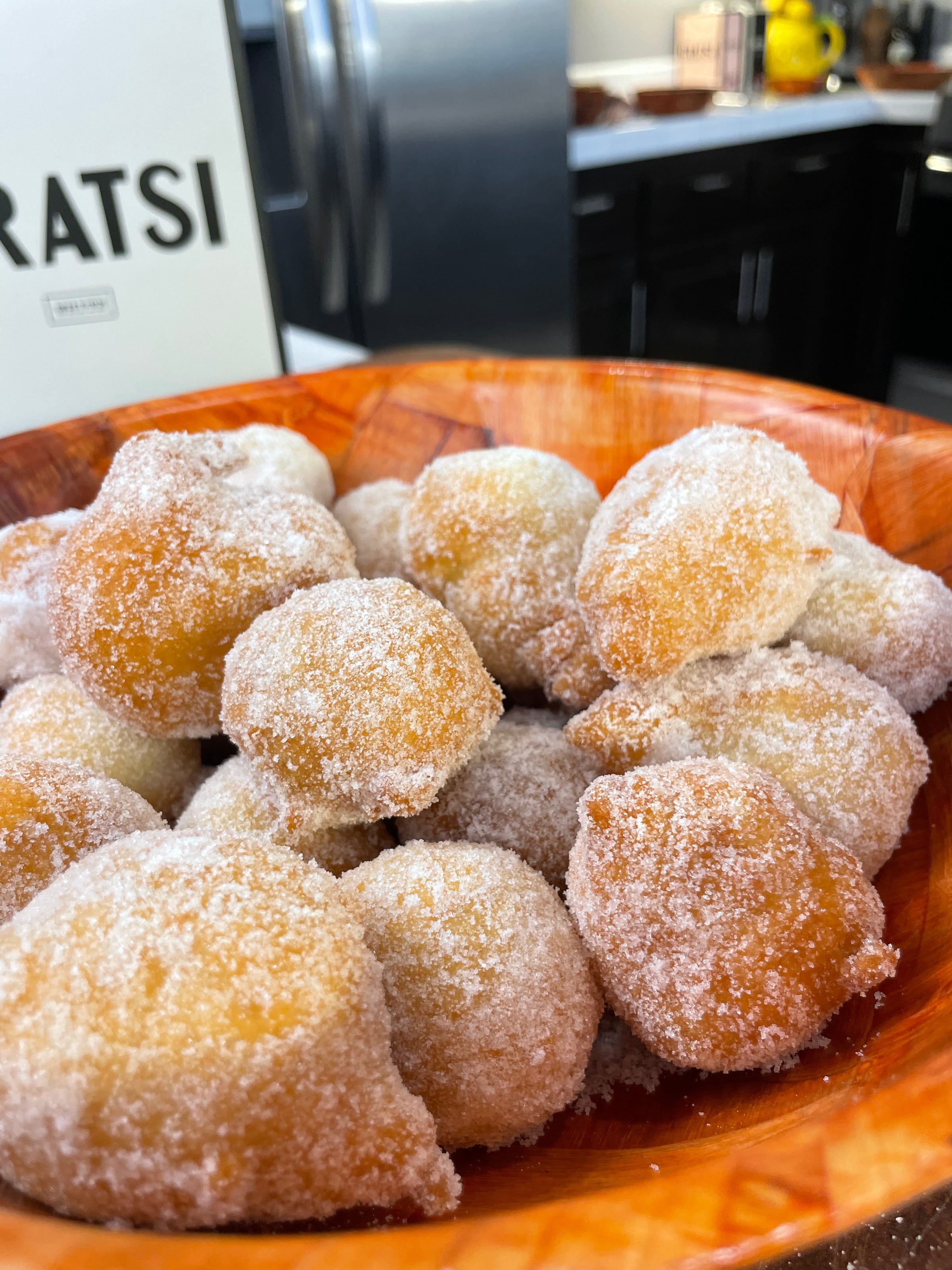 A wooden bowl filled with round, golden-brown doughnuts coated in sugar, placed on a kitchen counter with blurred cabinets and appliances in the background.