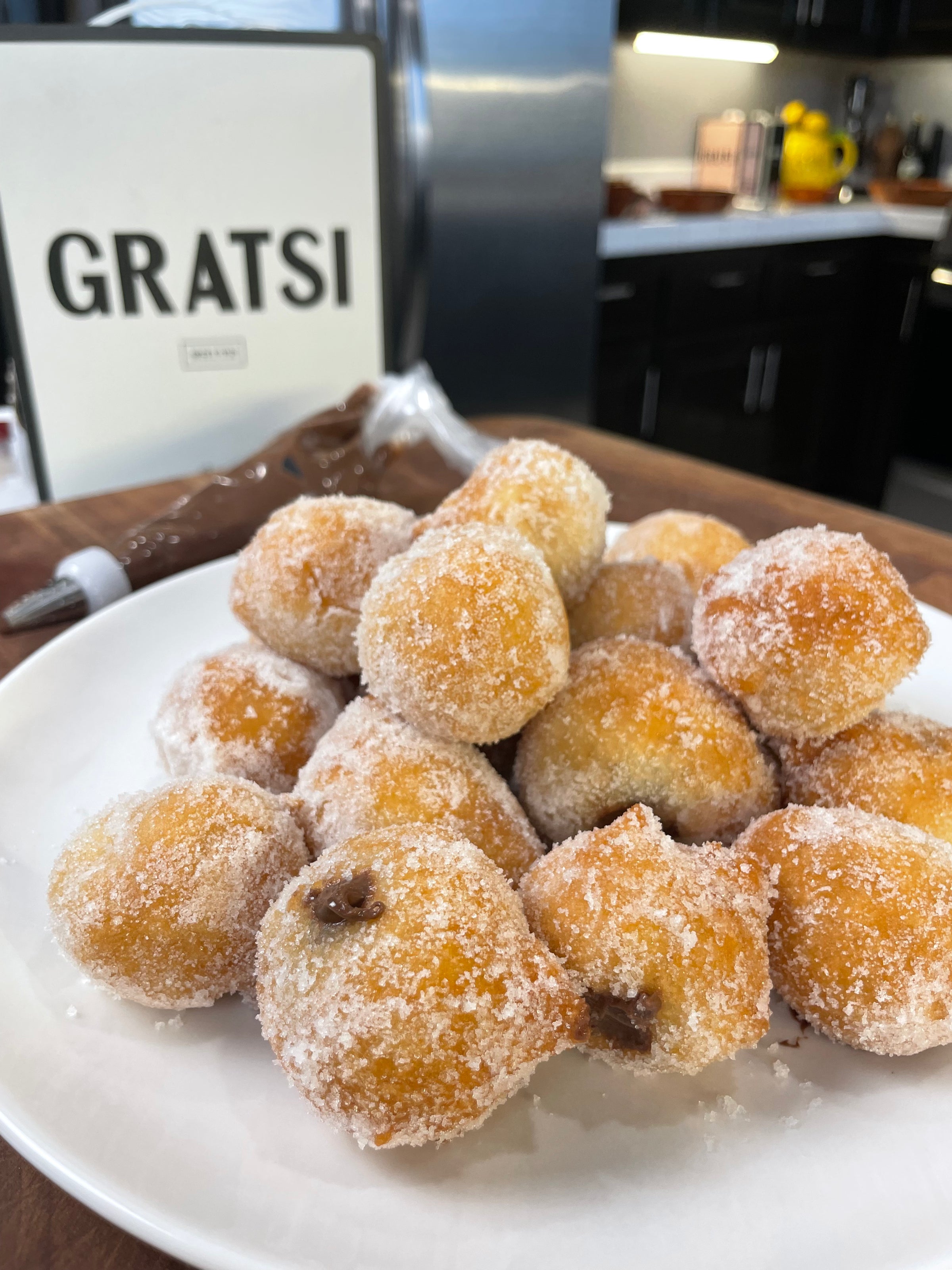 A plate of sugar-coated doughnut holes, some filled with chocolate, sits on a wooden counter in a kitchen. In the background, a sign reads GRATSI and a piping bag filled with chocolate is visible.